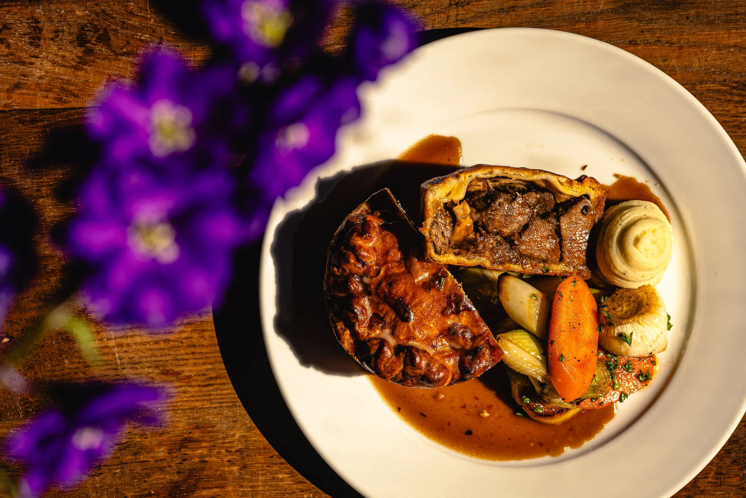 A white plate with slices of roast beef, roasted vegetables, and a swirl of mashed potatoes topped with parsley, served with brown gravy, on a wooden table, with purple flowers in the foreground.