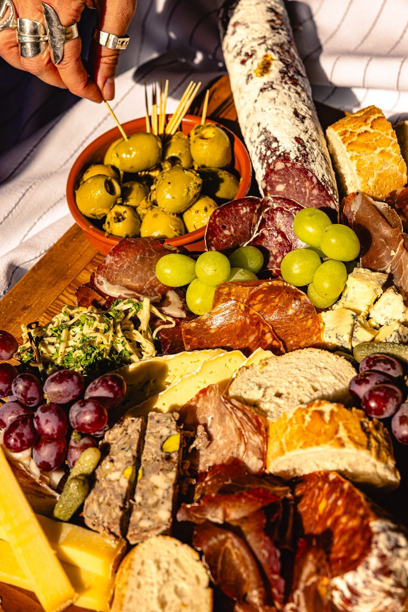 A cheese and charcuterie board with green olives, grapes, sliced cured meats, cheeses, crackers, and bread, served on a wooden platter.