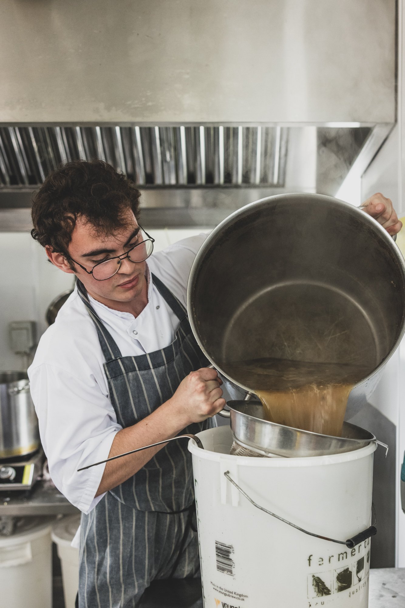 A young man with dark curly hair, glasses, and a striped apron is pouring a thick, brown liquid from a large metal container into a smaller one in a commercial kitchen.