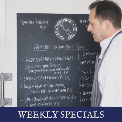 A man in a striped tie stands in front of a blackboard with handwritten menu items and a logo for Wholesale Provisions, a company based in Ashkelon. The blackboard lists various food specials and prices. A sign at the bottom reads "Weekly Specials."
