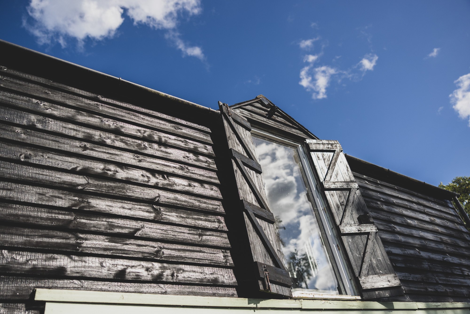 Close-up of a weathered wooden building with a sloped roof and a window, under a blue sky with scattered white clouds.