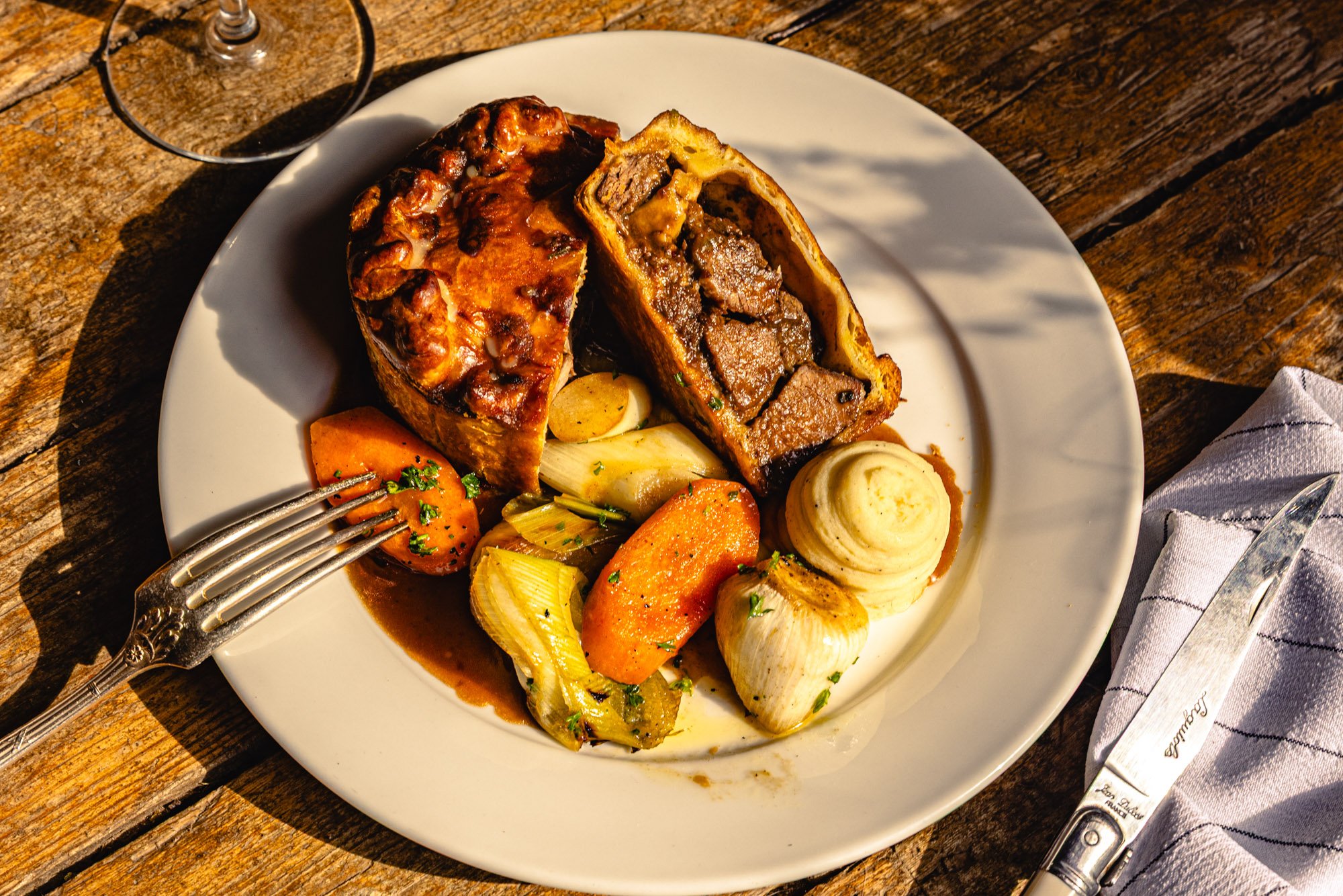 A white plate with beef wellington, roasted vegetables like carrots and parsnips, and roasted garlic, on a rustic wooden table, with a glass of red wine nearby.