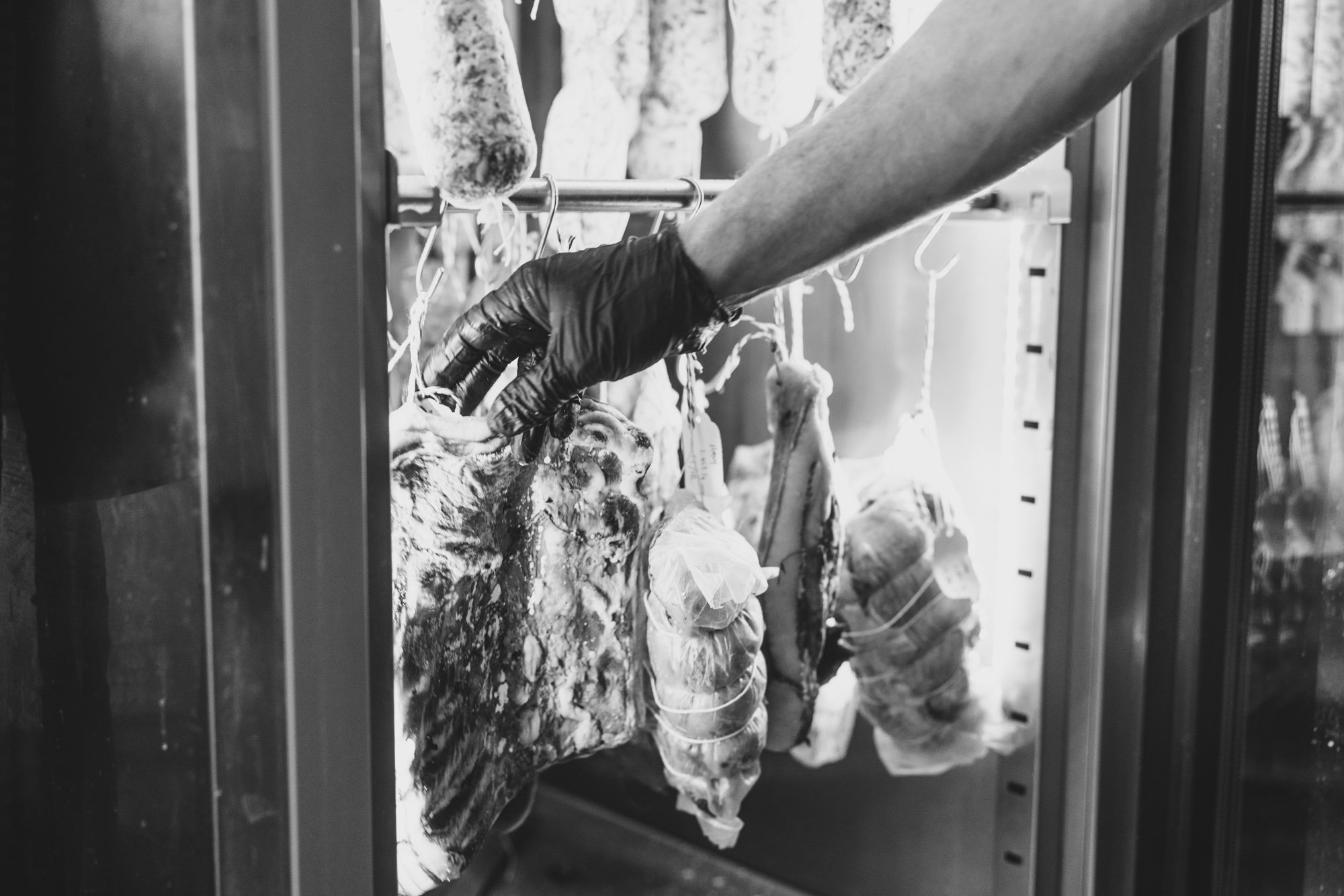 Black and white photo of hanging sausages and cured meats inside a refrigerated display case.