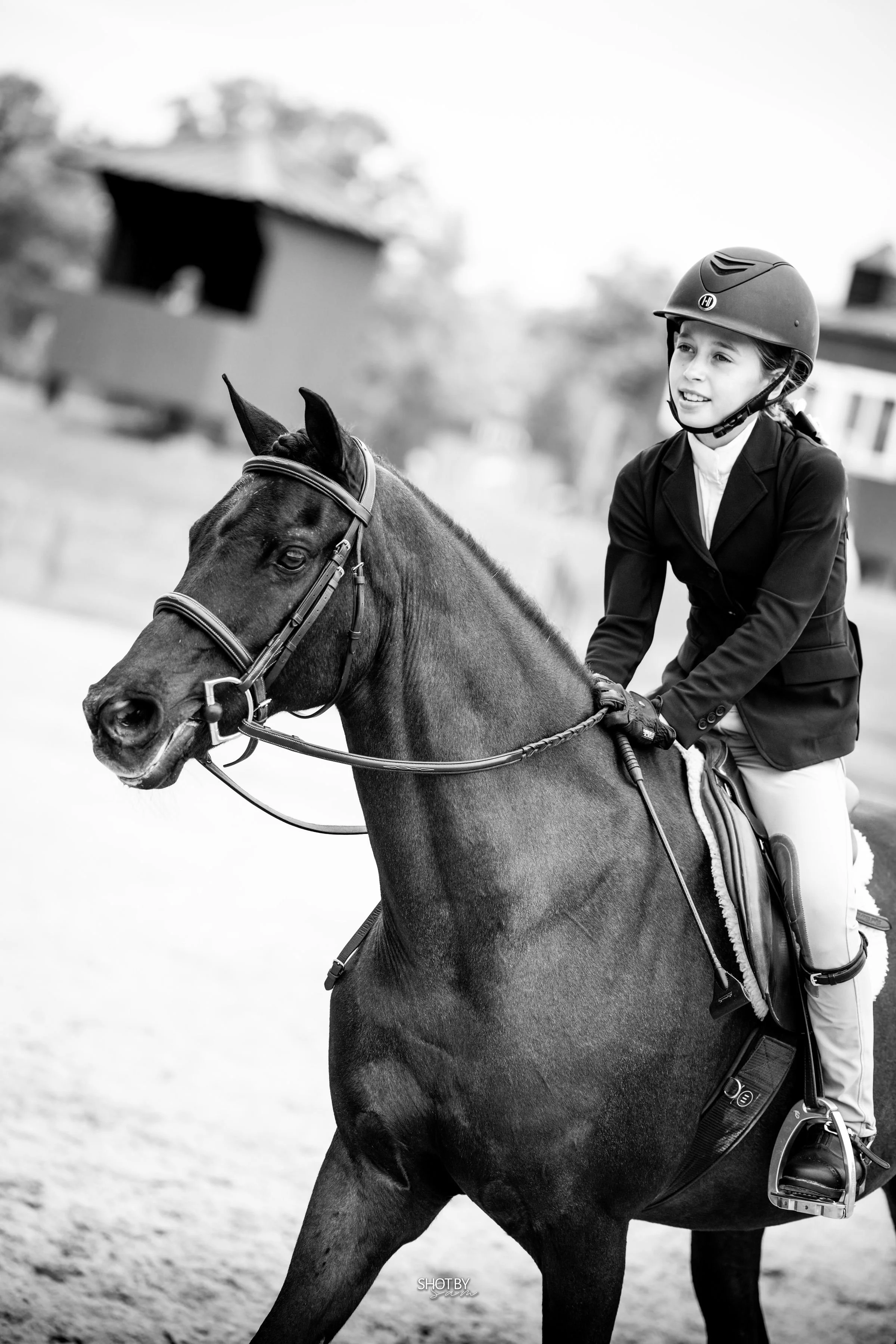 Young girl riding a horse, wearing riding gear including a helmet, in an outdoor setting.