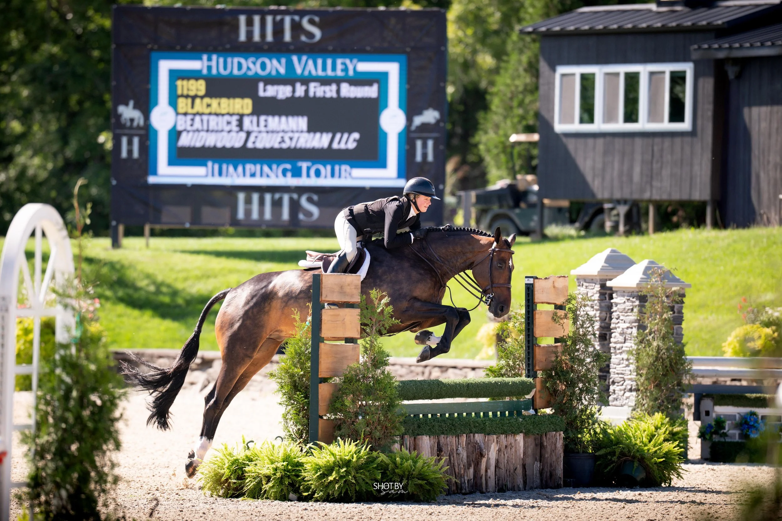 Equestrian rider on a horse jumping over an obstacle during a competition at Hudson Valley. Background includes large sign, building, and greenery.