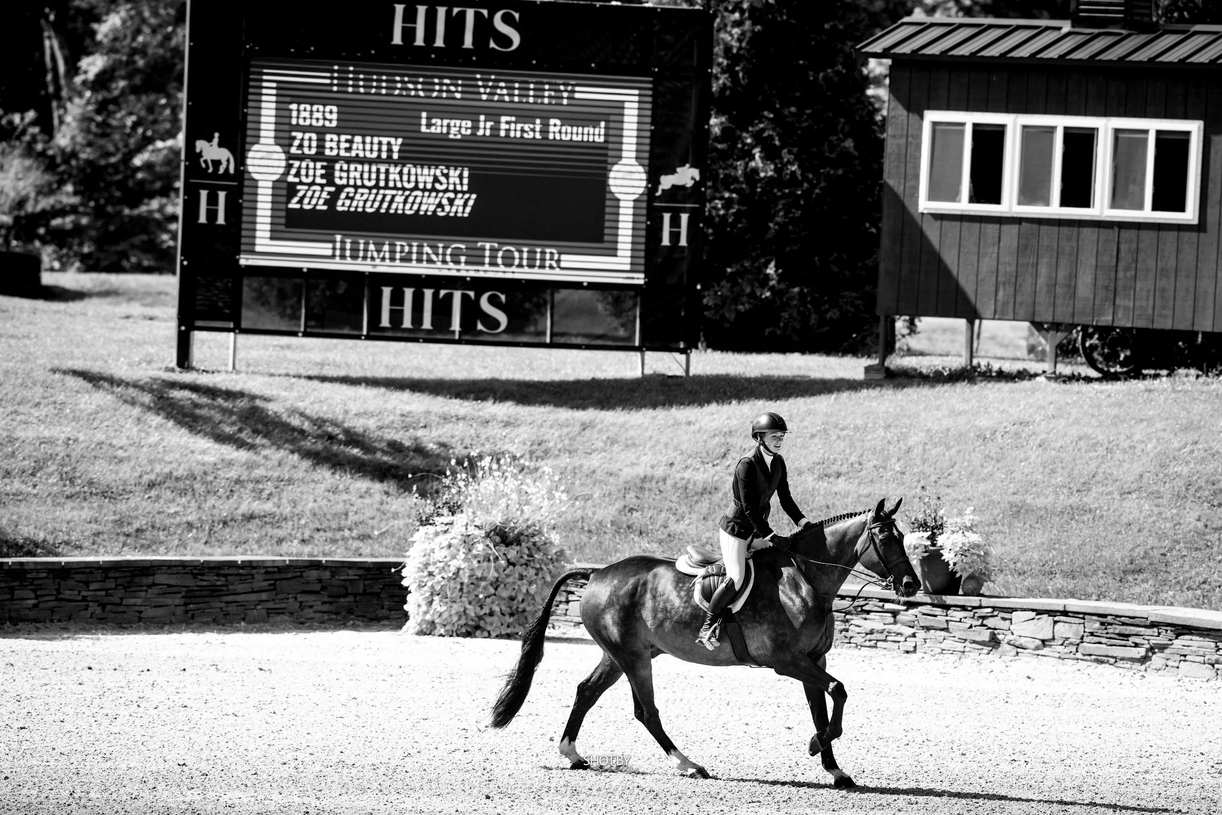 A person riding a horse in an outdoor riding arena, with a large sign on the side displaying horse jumping event details and a small building with windows in the background.