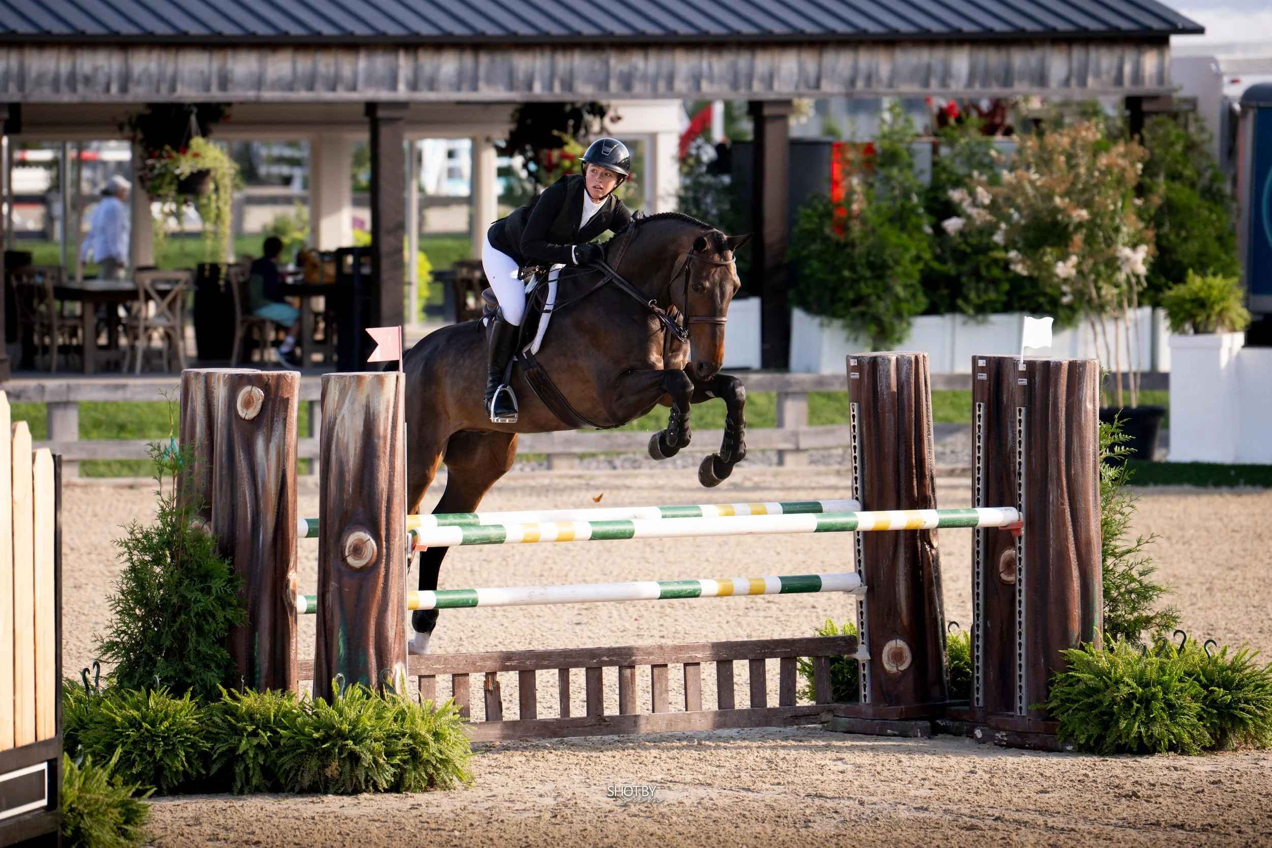 A female equestrian rider in formal riding attire and a helmet jumping her horse over a show jump at an outdoor equestrian competition.