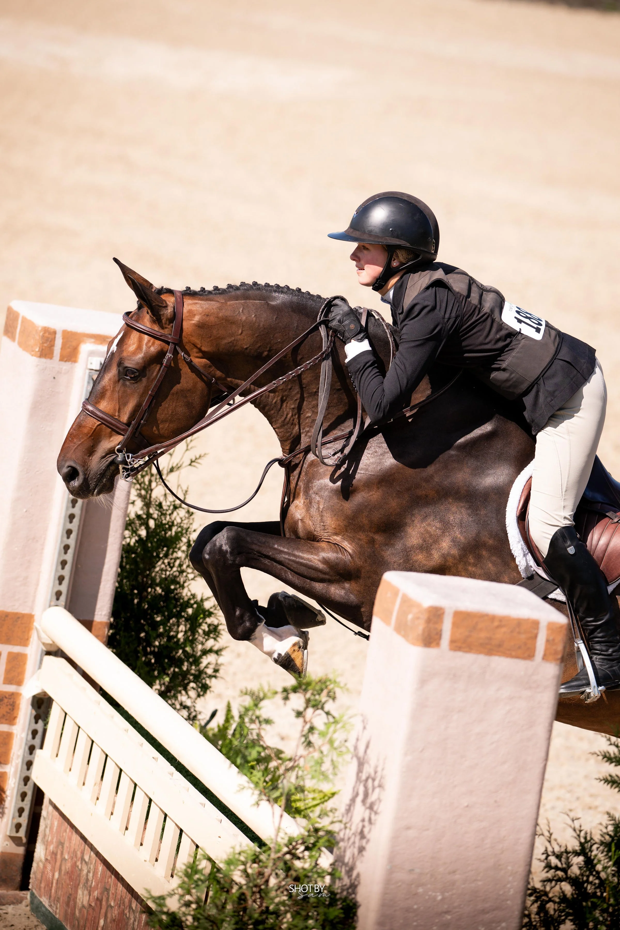 A female equestrian athlete wearing a black helmet and riding gear navigates a jump during a show jumping competition, in an outdoor arena with a beige wall background.