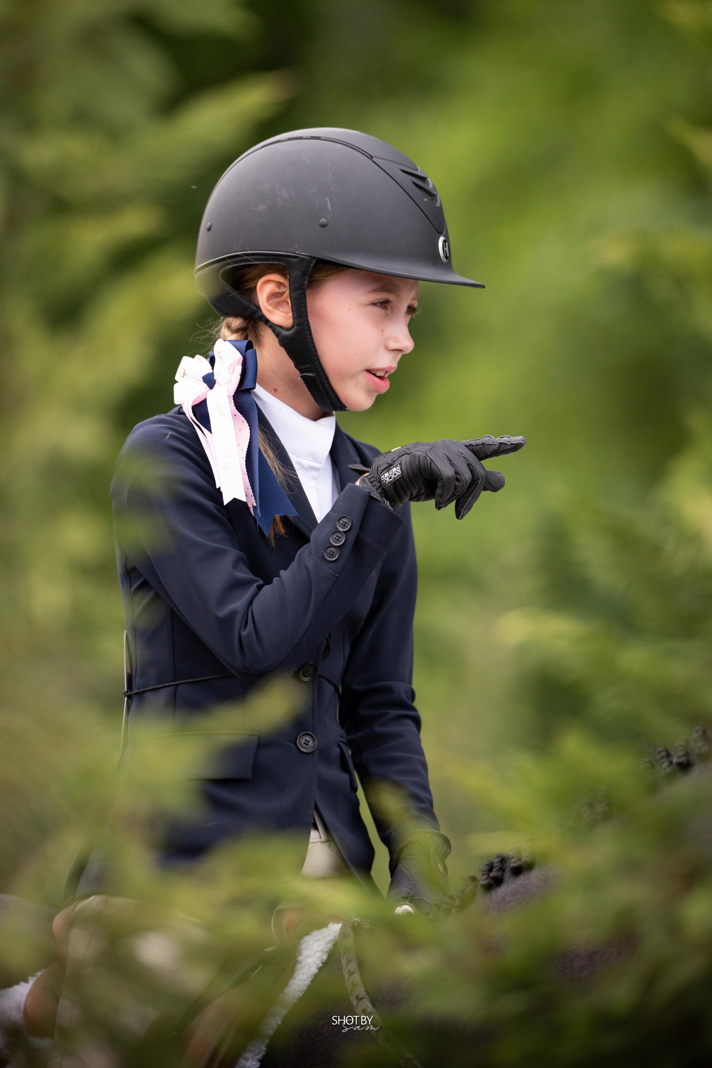 A young girl in equestrian riding gear, including a black helmet, navy jacket, and gloves, riding a horse through a green outdoor setting.