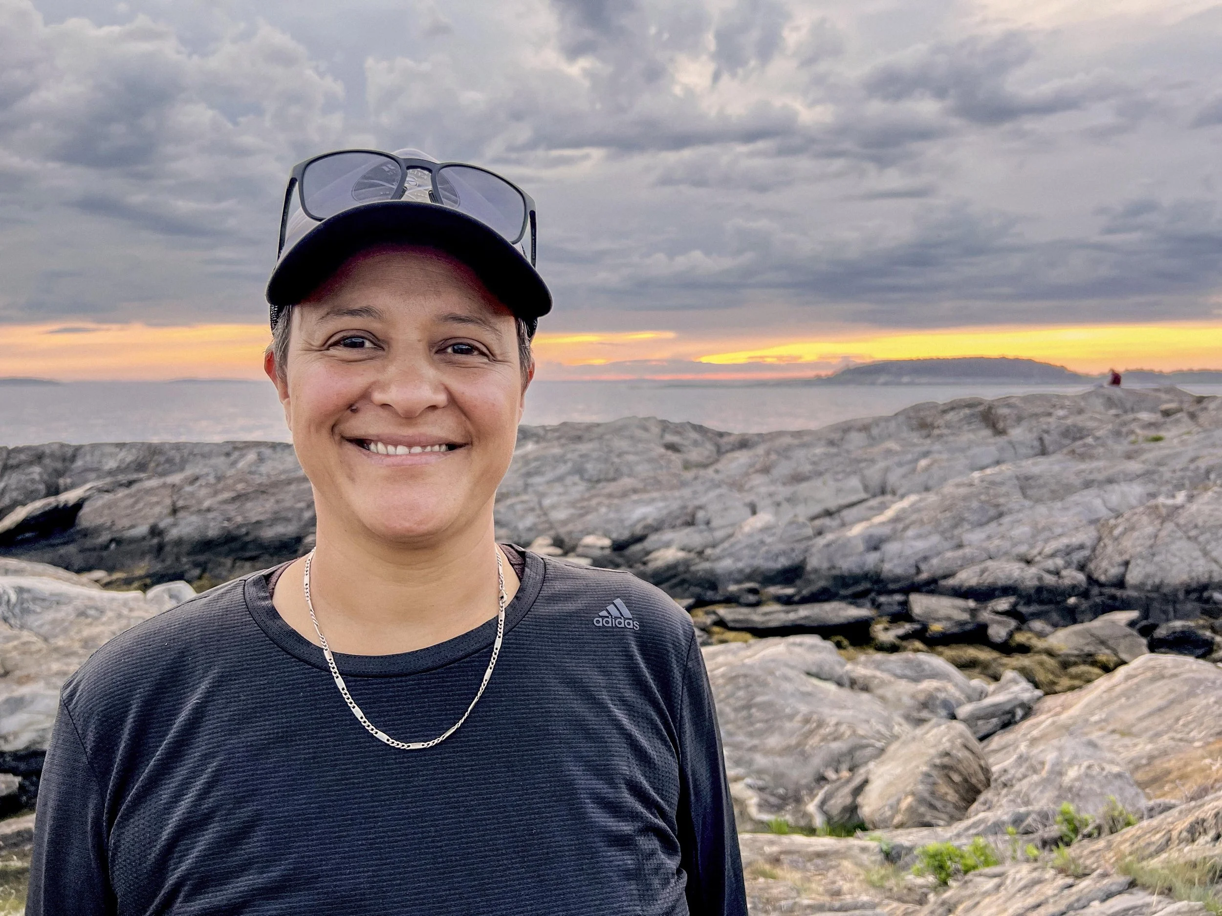A smiling woman wearing a black Adidas athletic shirt, a black cap with sunglasses resting on top, standing on a rocky shoreline at sunset with a cloudy sky and distant islands in the background.
