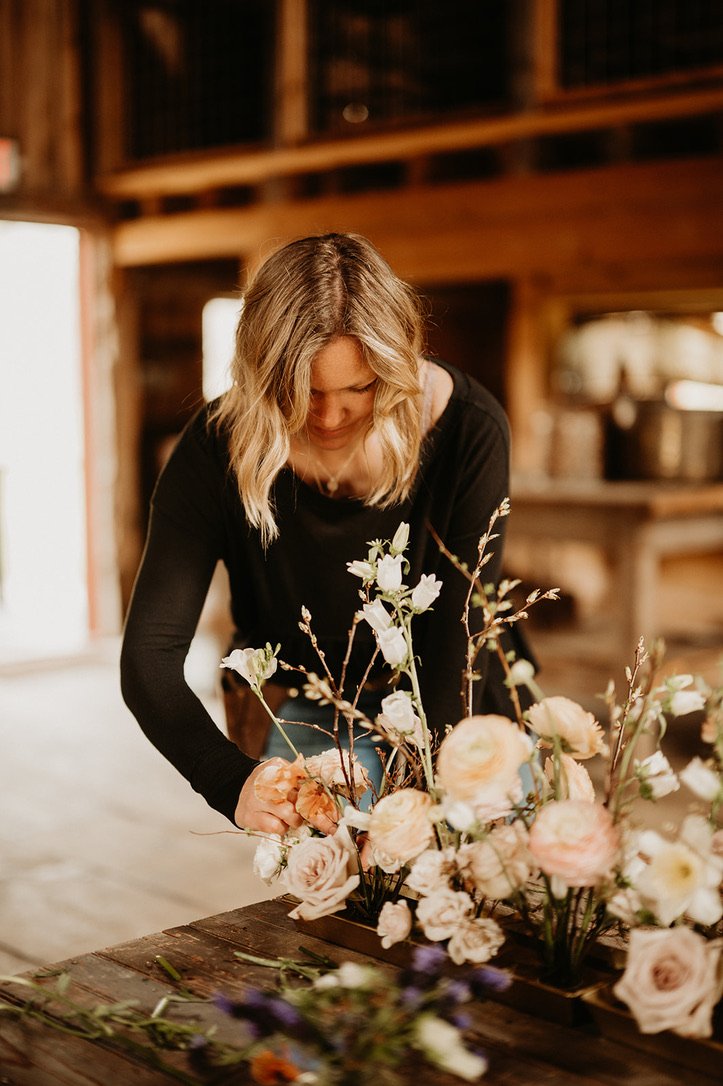 Lindsay, our floral designer, arranging a bouquet of flowers on a rustic wooden table in a cozy, wooden interior, photograph taken by wedding photographer, Katelyn Mallet.