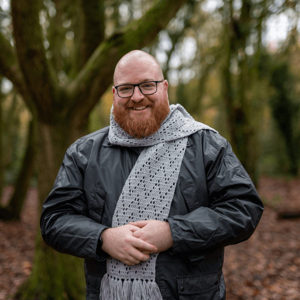 bald man with a ginger beard wearing a black coat and a grey crocheted scatf with a zigzag motif.png
