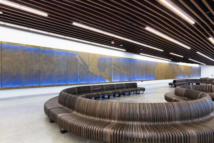 Visual Voyage, a modern cruise ship waiting area like an airport terminal with curved wooden benches and a large world map on a backlit wall. This interactive installation features programmable lighting and interactive via personal devices.