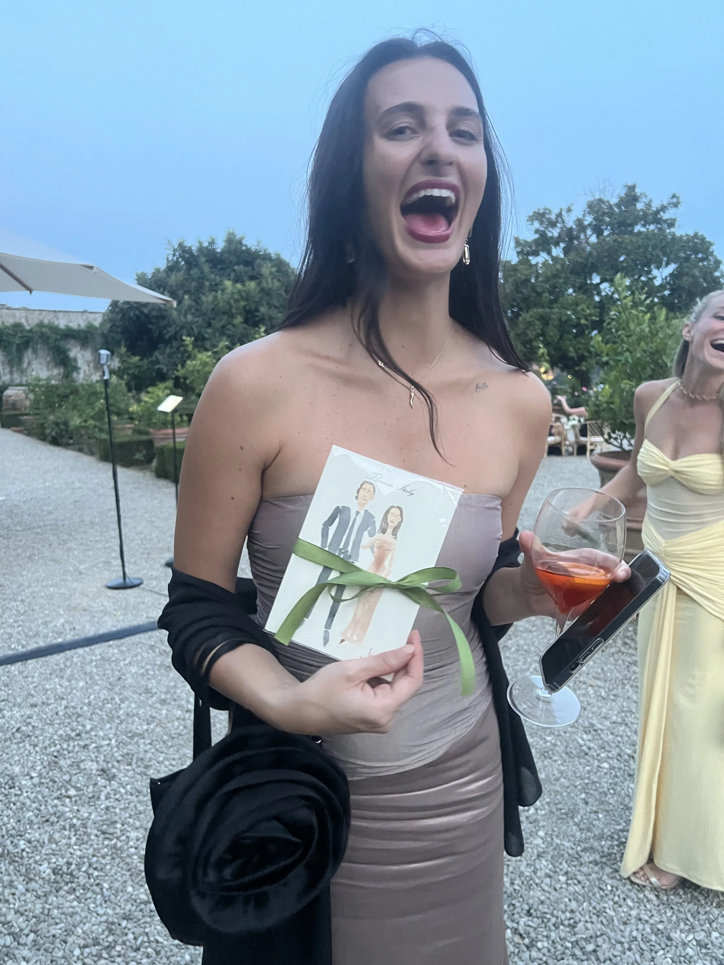 A woman with long dark hair smiling and holding an Italy wedding favor, a glass of pink drink, and a smartphone, at  Villa Corsini, a Tuscan wedding in the evening with other women in the background.