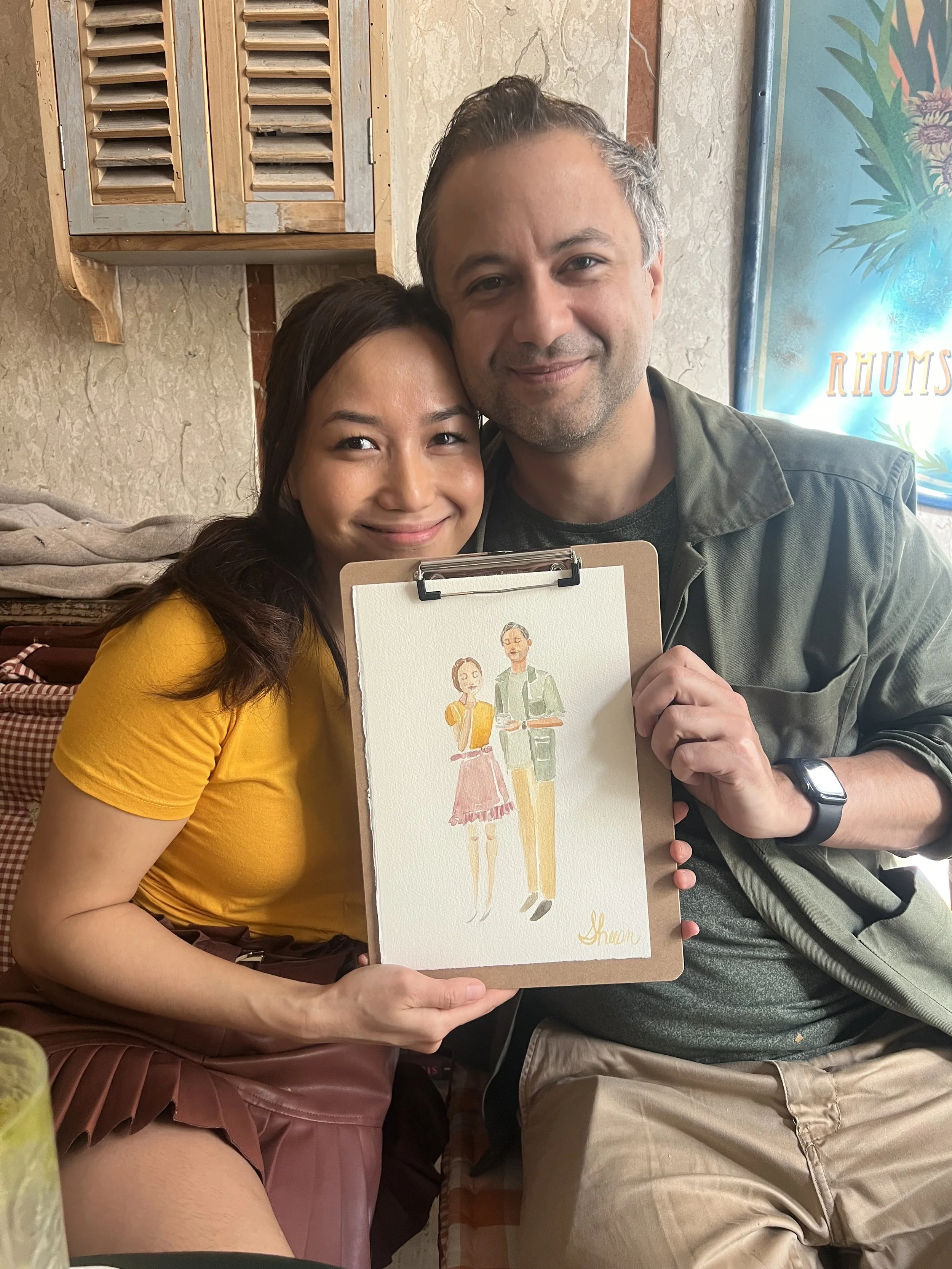 A smiling woman and man sitting together inside a cafe, holding a hand-painted Florence souvenir of a woman and man. The woman in the painting is wearing a pink skirt and yellow top, and the man is wearing yellow pants and a green jacket. The woman i