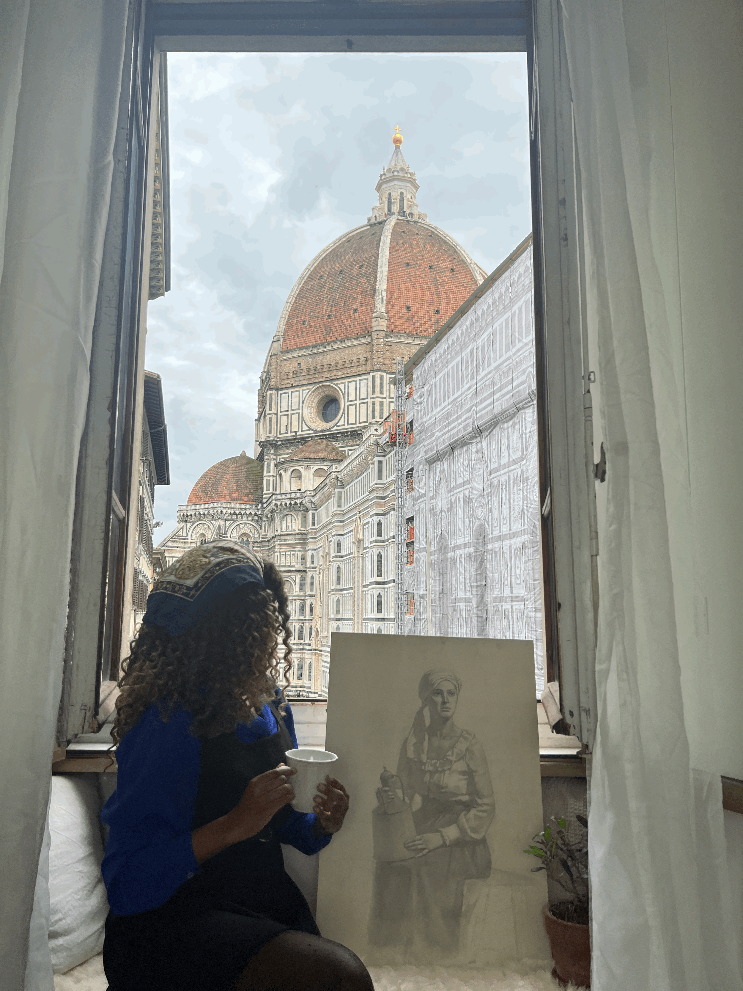 A woman with curly hair, wearing a headscarf and a blue and black jacket, sitting by a window with a view of Florence Cathedral's dome, holding a cup. There is a portrait of a woman with a lamp, and a potted plant on the right.
