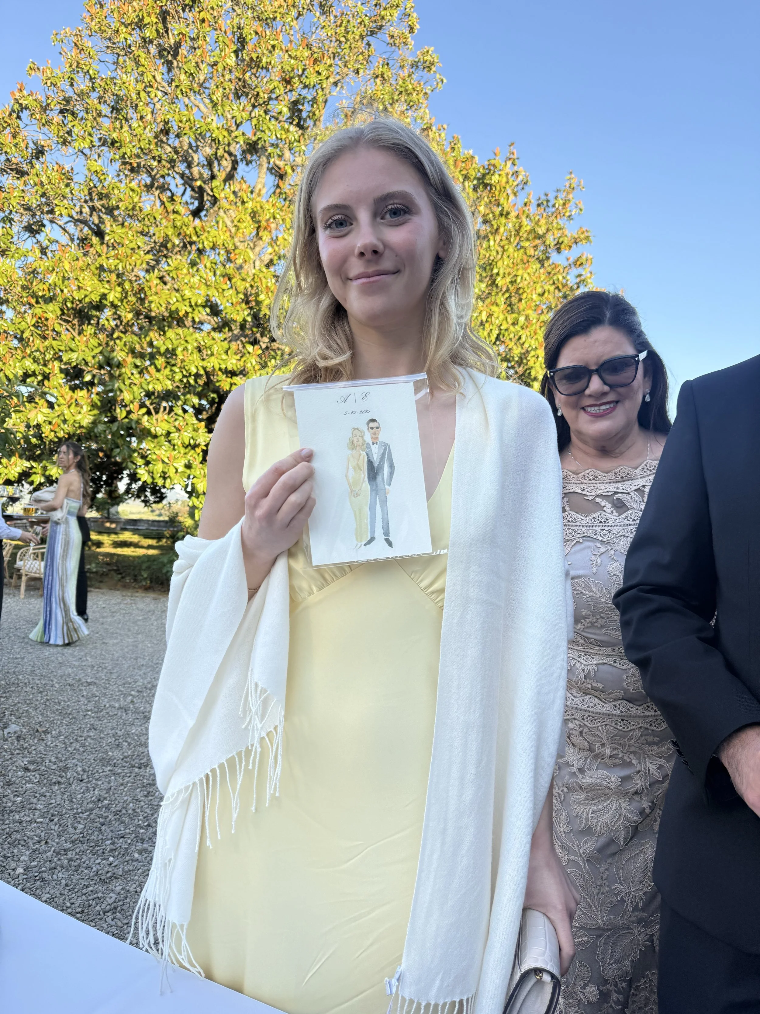 A young woman in a light yellow dress holding a hand-drawn Italy live wedding guest portrait of a couple, standing at Villa Corsini near a large tree with green leaves under a clear blue sky, with other people visible in the background.