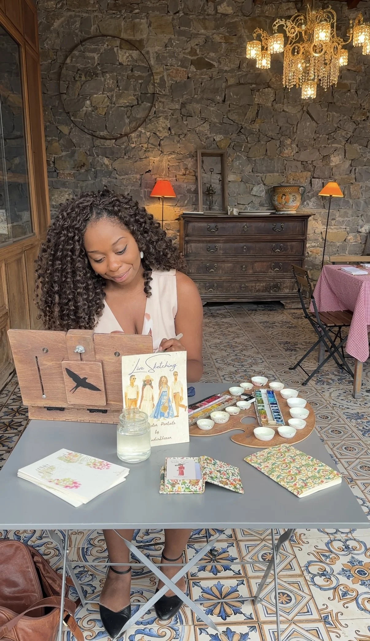 Artist Studio Sheean live-sketching watercolor guest portraits sitting at a table painting fashion illustrations with watercolor paints. The table has decorative paper napkins, a glass of water, and sketches. The background features a stone wall, a c