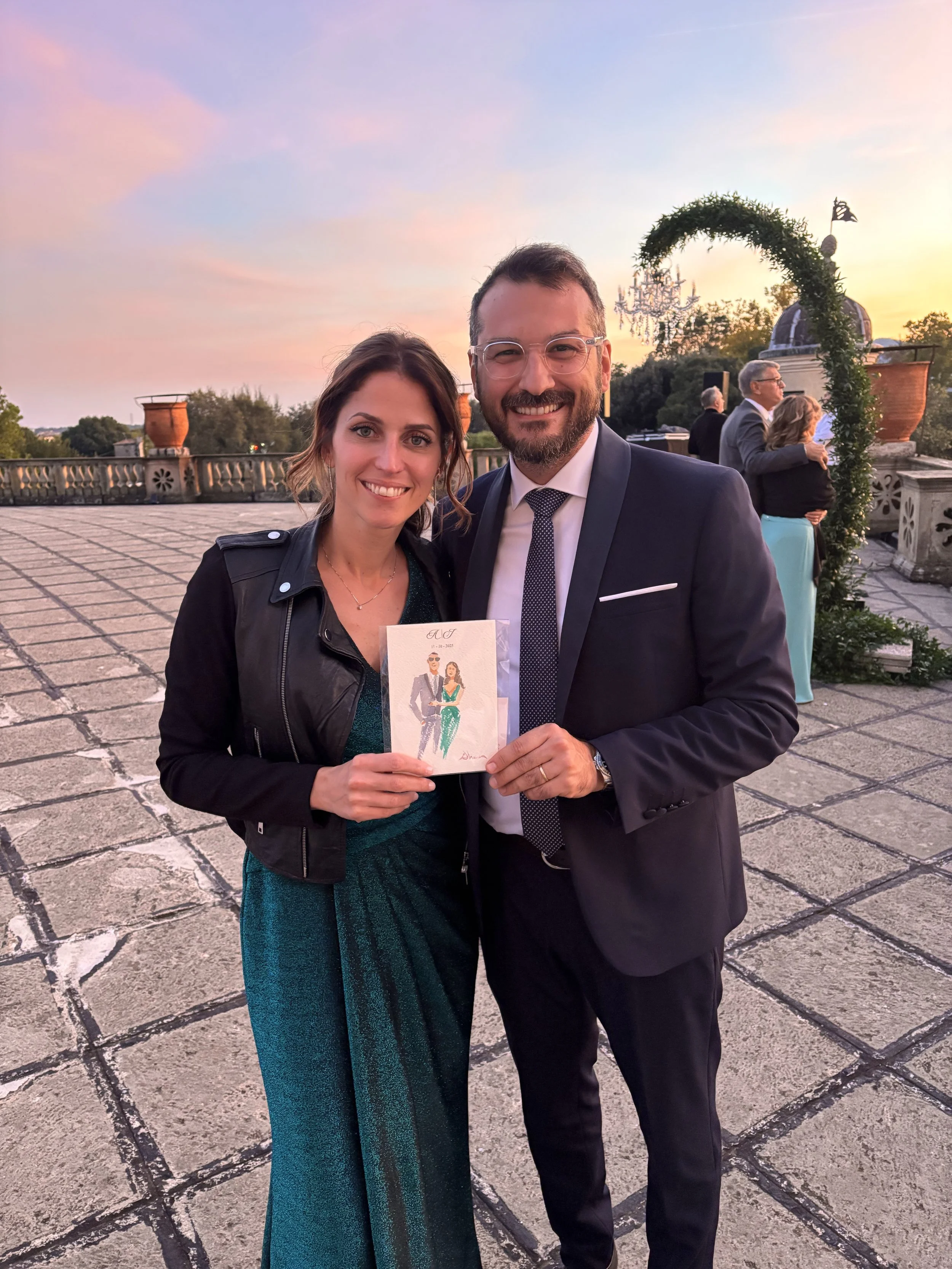 A smiling woman and man in formal attire standing outside at sunset. The woman holds a unique Italy wedding favor painted in watercolor.