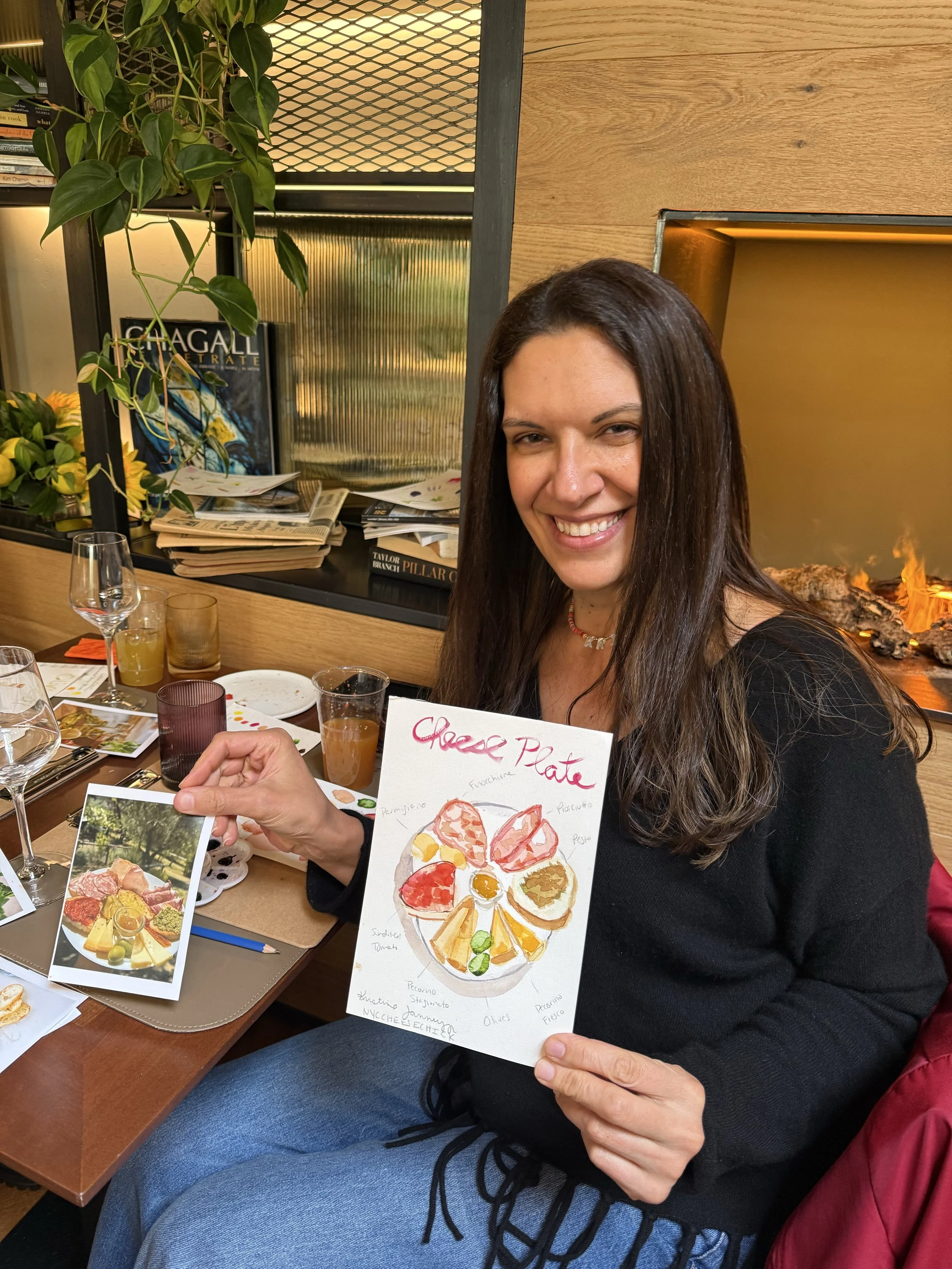 Woman smiling at the camera, holding a hand-drawn diagram of a cheese plate that labels various cheeses and accompaniments, seated at a restaurant table with various drinks and a photograph of a cheese platter on the table at a  watercolor painting w