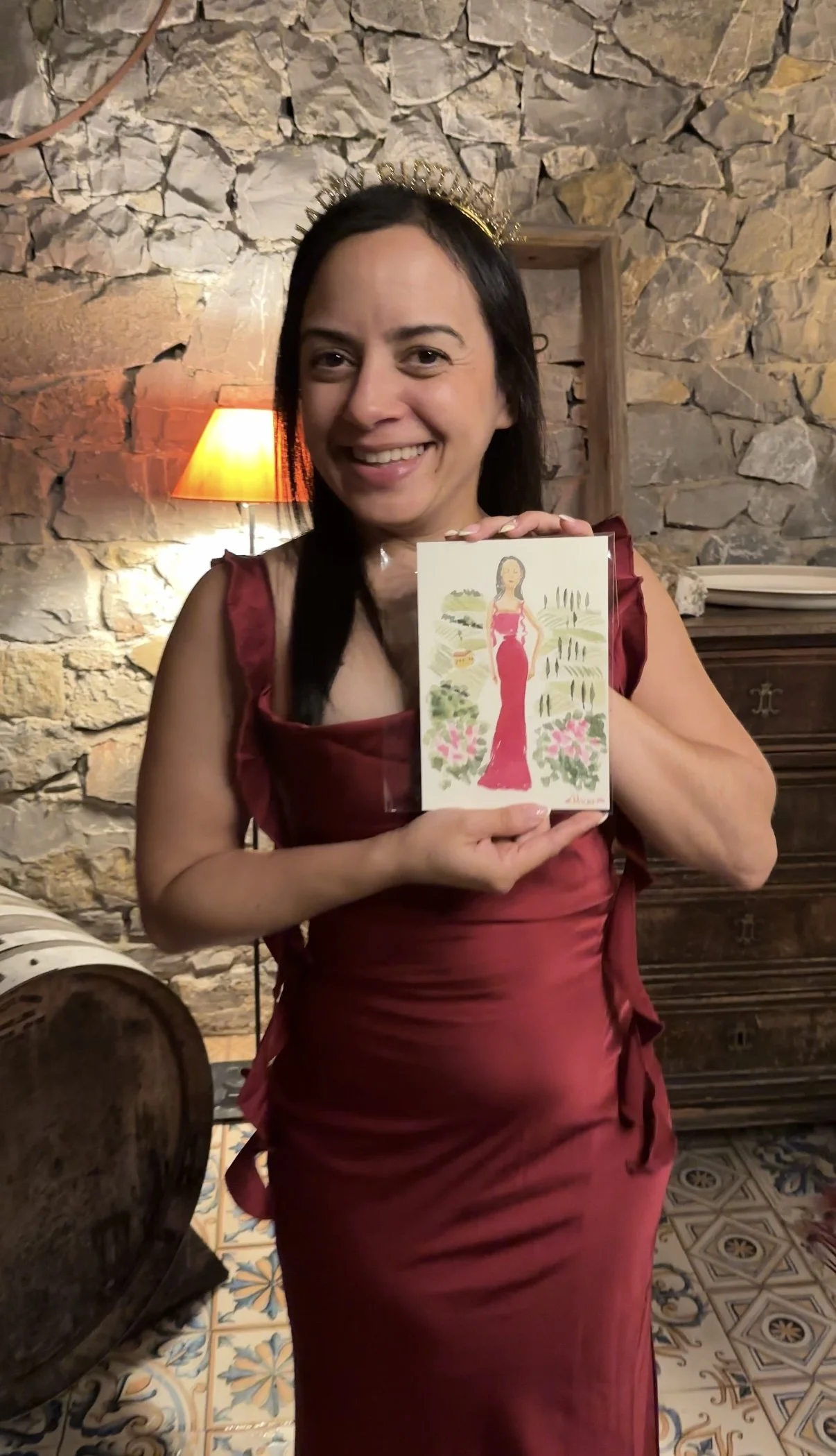 Woman smiling and wearing a crown, holding a Florence Italy watercolor souvenir featuring a woman in a red dress, in a room with stone walls and vintage furniture.