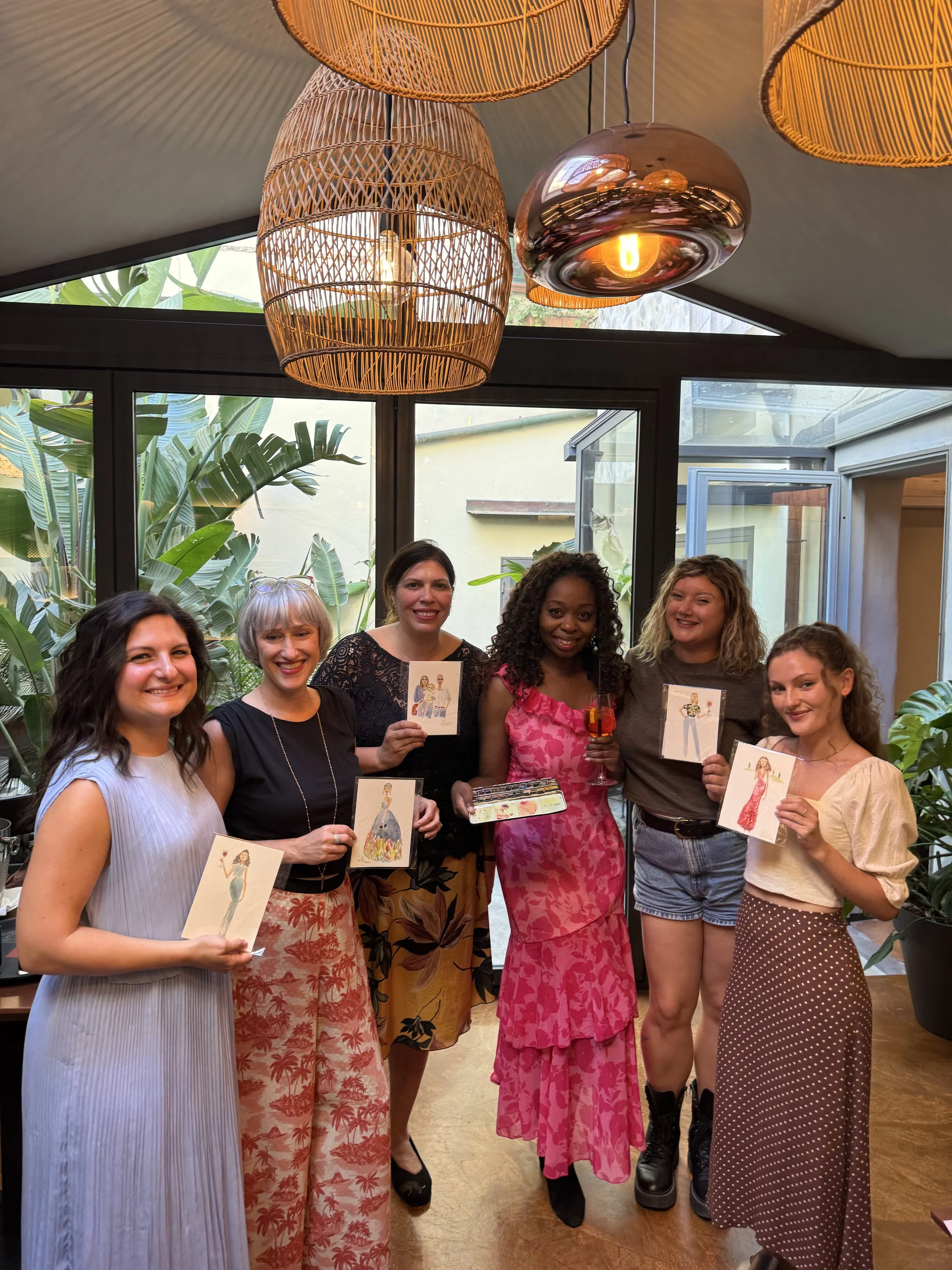 A diverse group of six women standing indoors, holding colorful Italy guest portraits illustrations of women in fashionable outfits, smiling at the camera, with lush greenery outside the windows behind them.