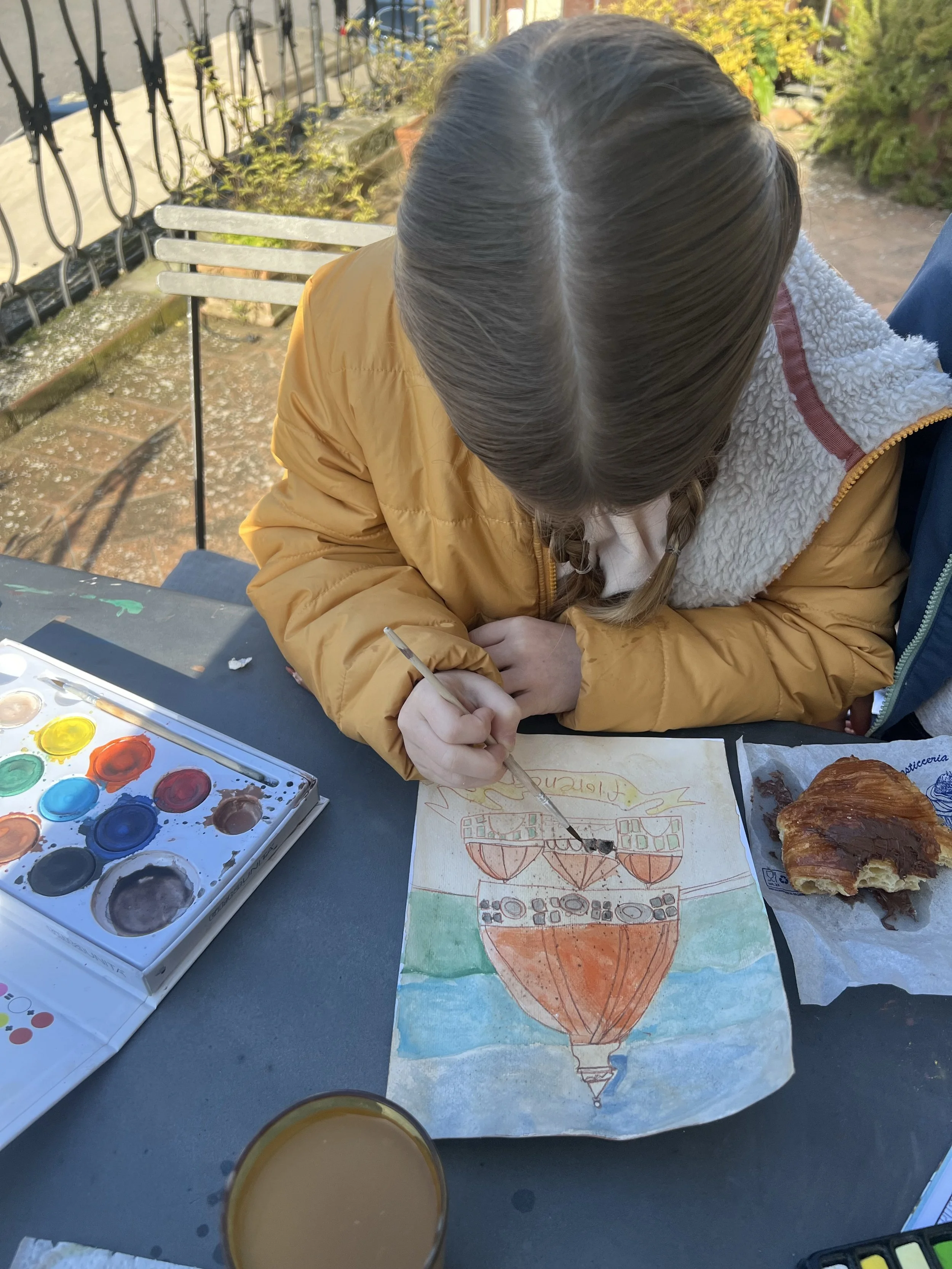 A girl in a yellow jacket is painting a picture of a hot air balloon, with watercolors and a paintbrush. There is a partially eaten croissant and a glass of coffee on the table.