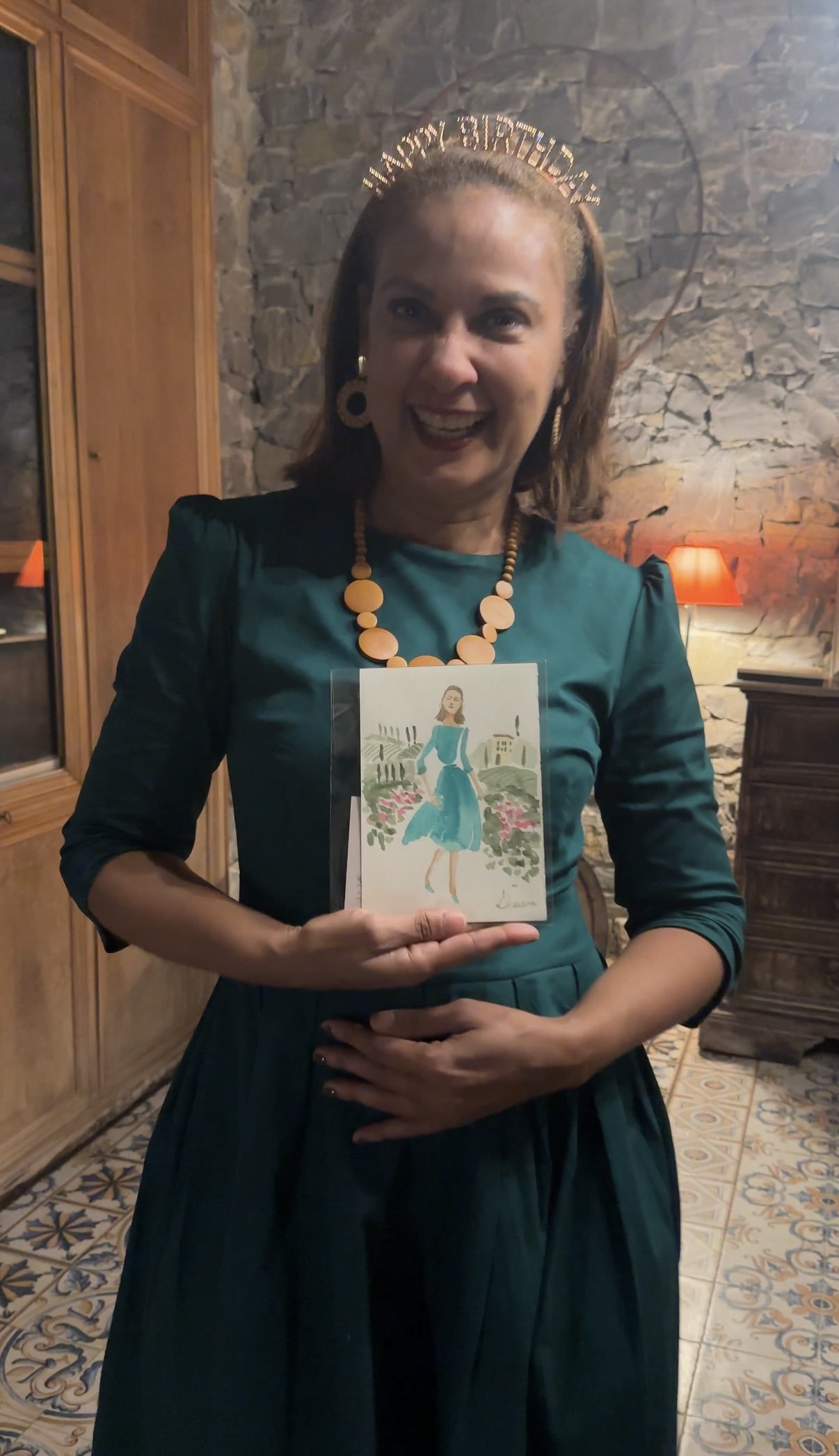 A woman smiling and holding a hand-painted watercolor souvenir in Florence, Italy in a green dress, with a stone wall and lamps in the background.