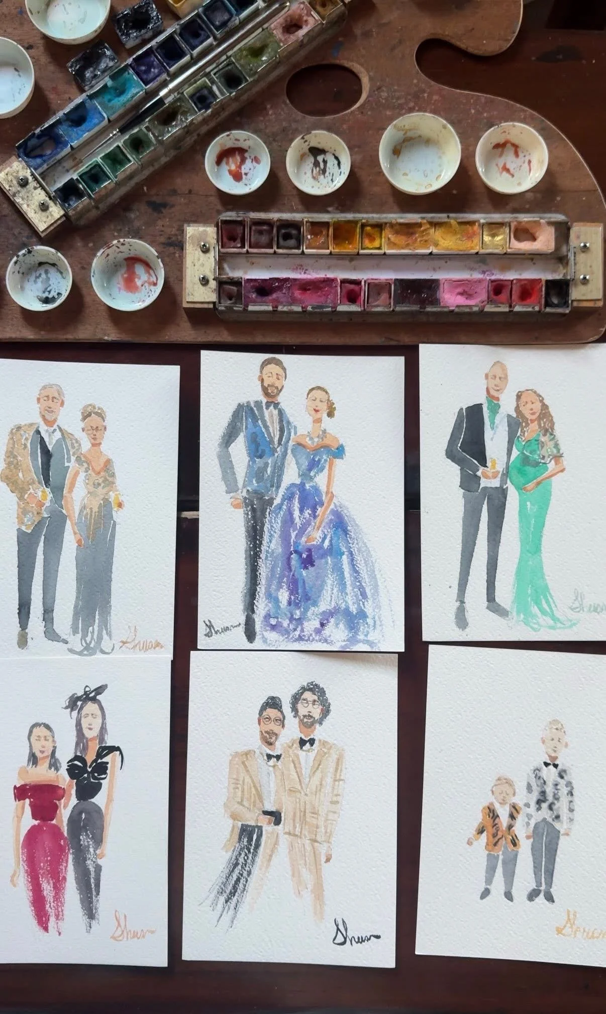 Italy live wedding watercolor paintings of various couples dressed in formal attire, laid out on a wooden table, with watercolor palettes and brushes at the top of the image.