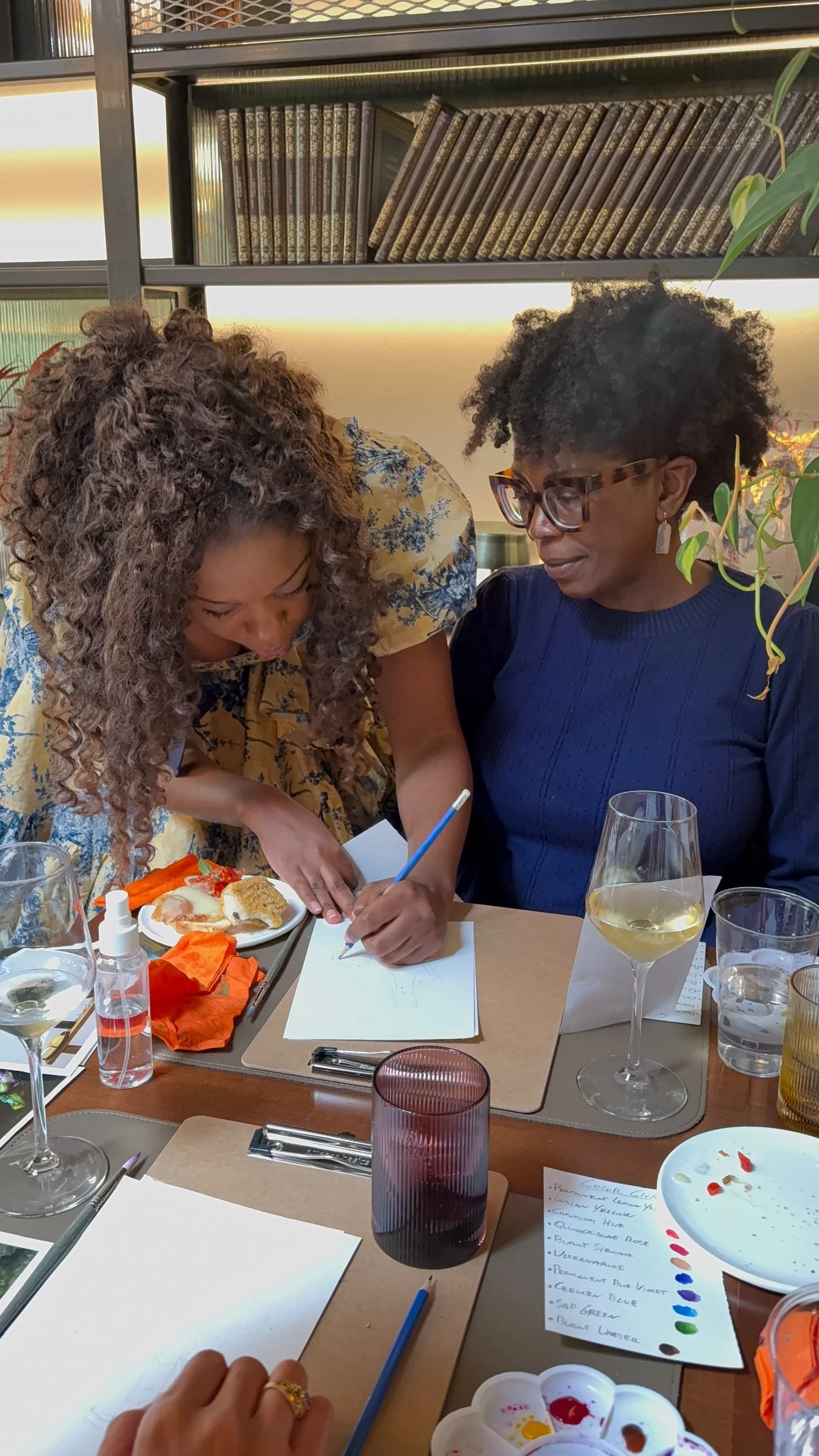 Two women are seated at a table, one is sketching and the other is observing. There are various art supplies, glasses of white wine, and plates of food on the table.