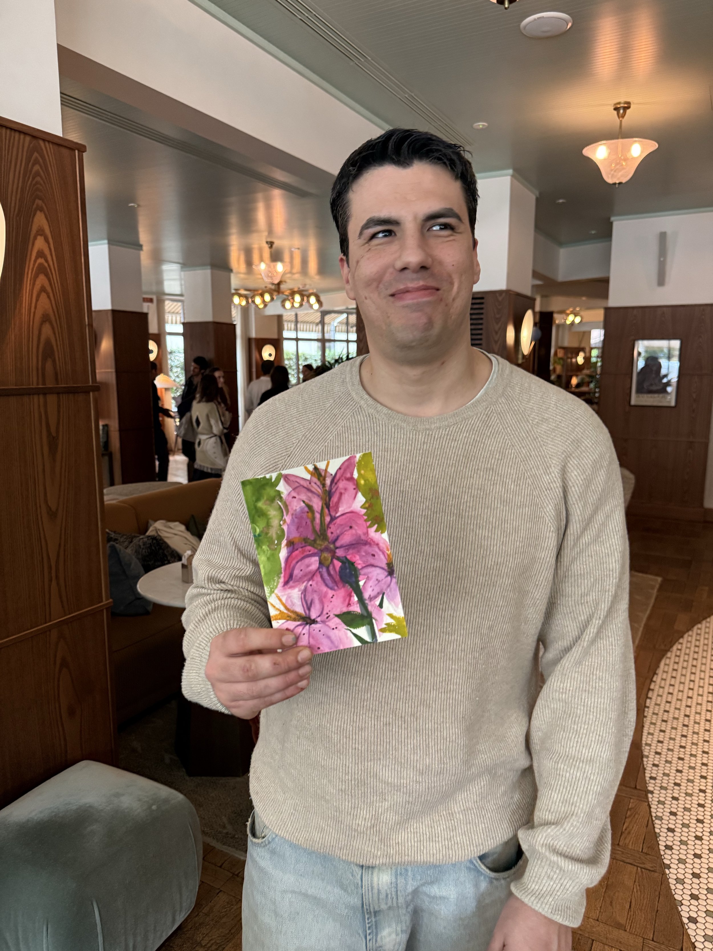 A man in a beige sweater holding a colorful painting of pink flowers with green leaves, standing inside a warmly lit restaurant or cafe.