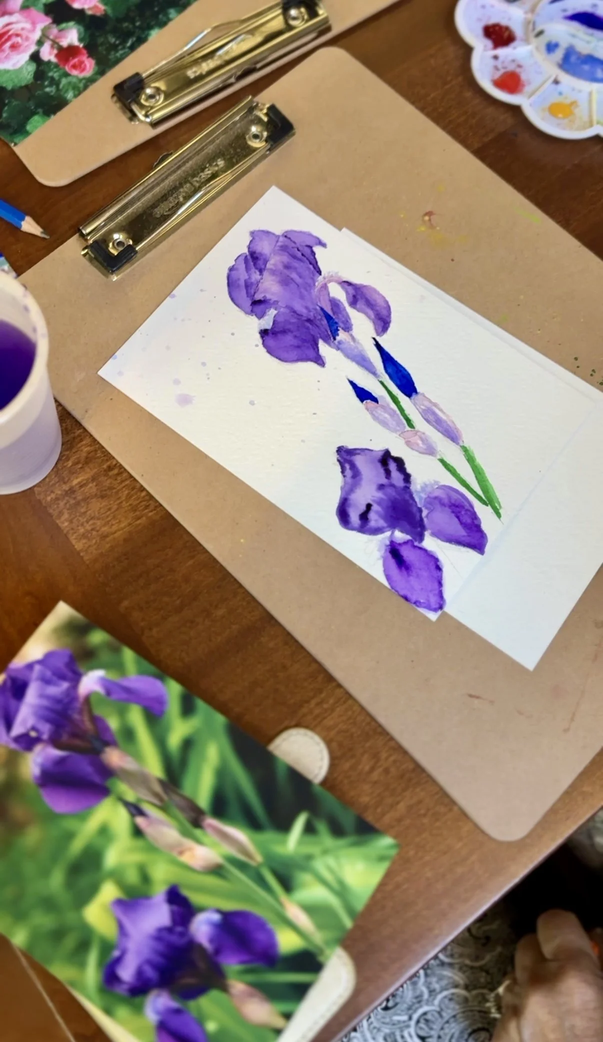 Watercolor painting of purple irises on white paper, with reference photo of real irises nearby, on a wooden table with painting supplies around during a private art lesson in Florence Italy.