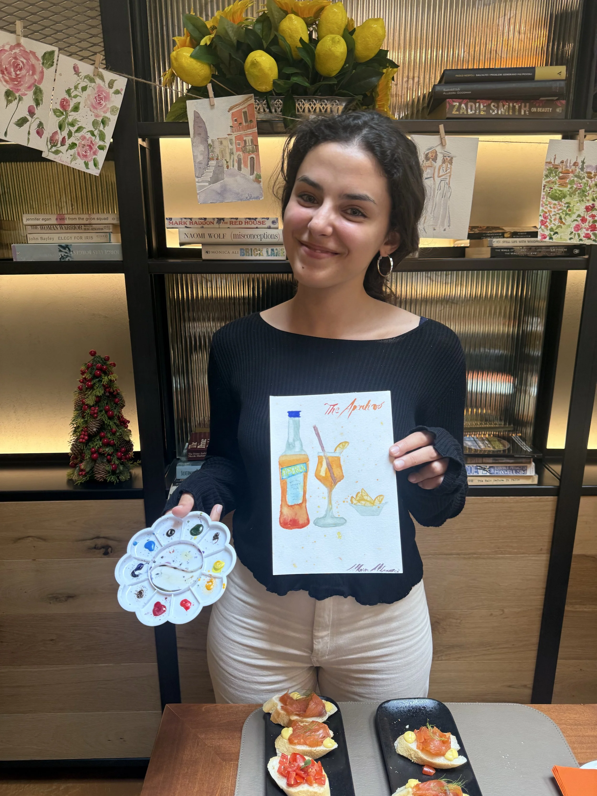A woman smiling inside a room decorated with books, artwork, and yellow lemons on a vase, holding a painting of a cocktail with a bottle, glass, lemon wedge, and ice. In front of her on a table are three small appetizers topped with diced tomatoes, s