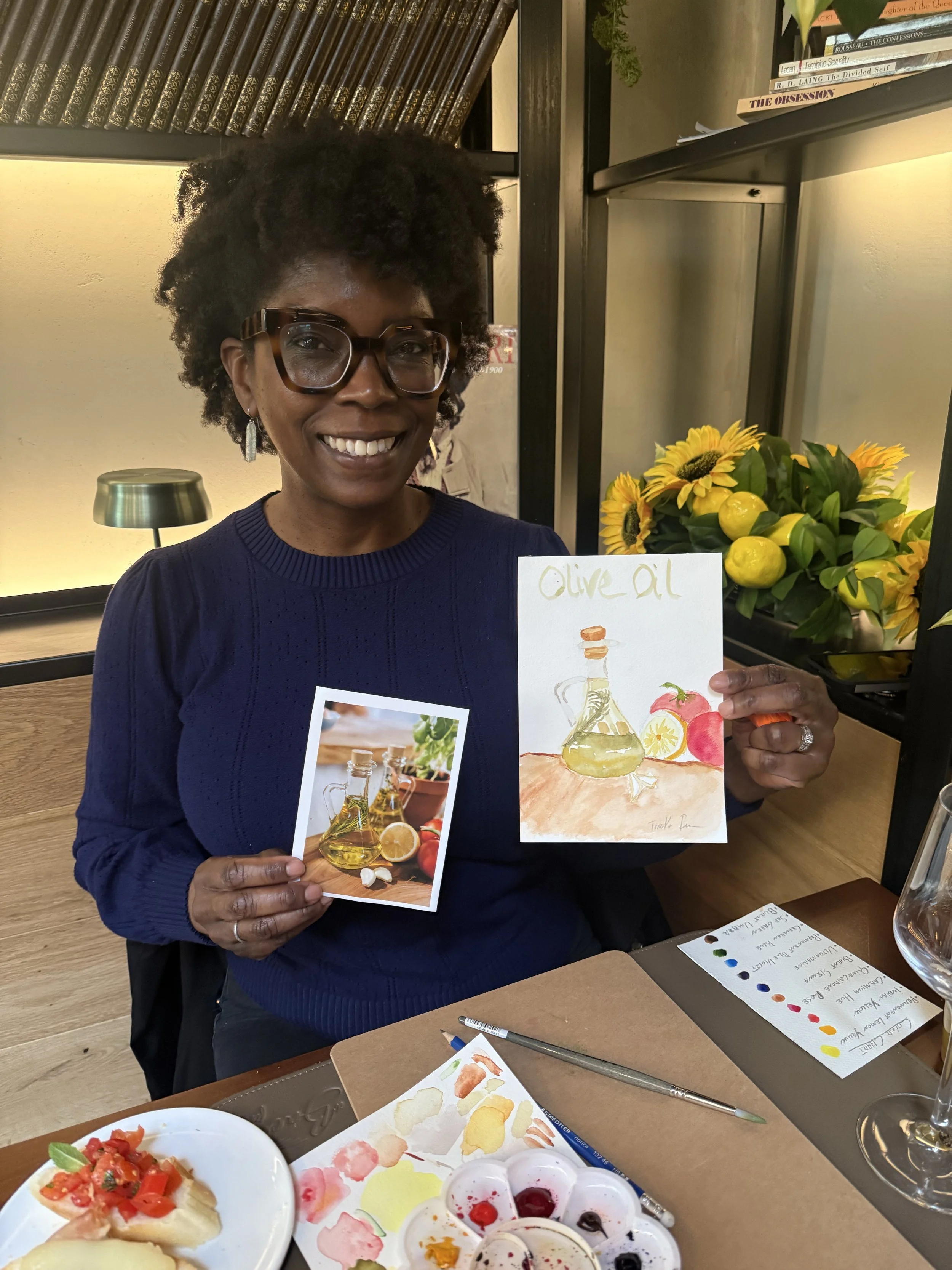A smiling woman with glasses, short curly hair, in a blue sweater, holding a card with a watercolor painting of olive oil. Bespoke live sketching for tourists looking for authentic art experiences in Florence Italy.