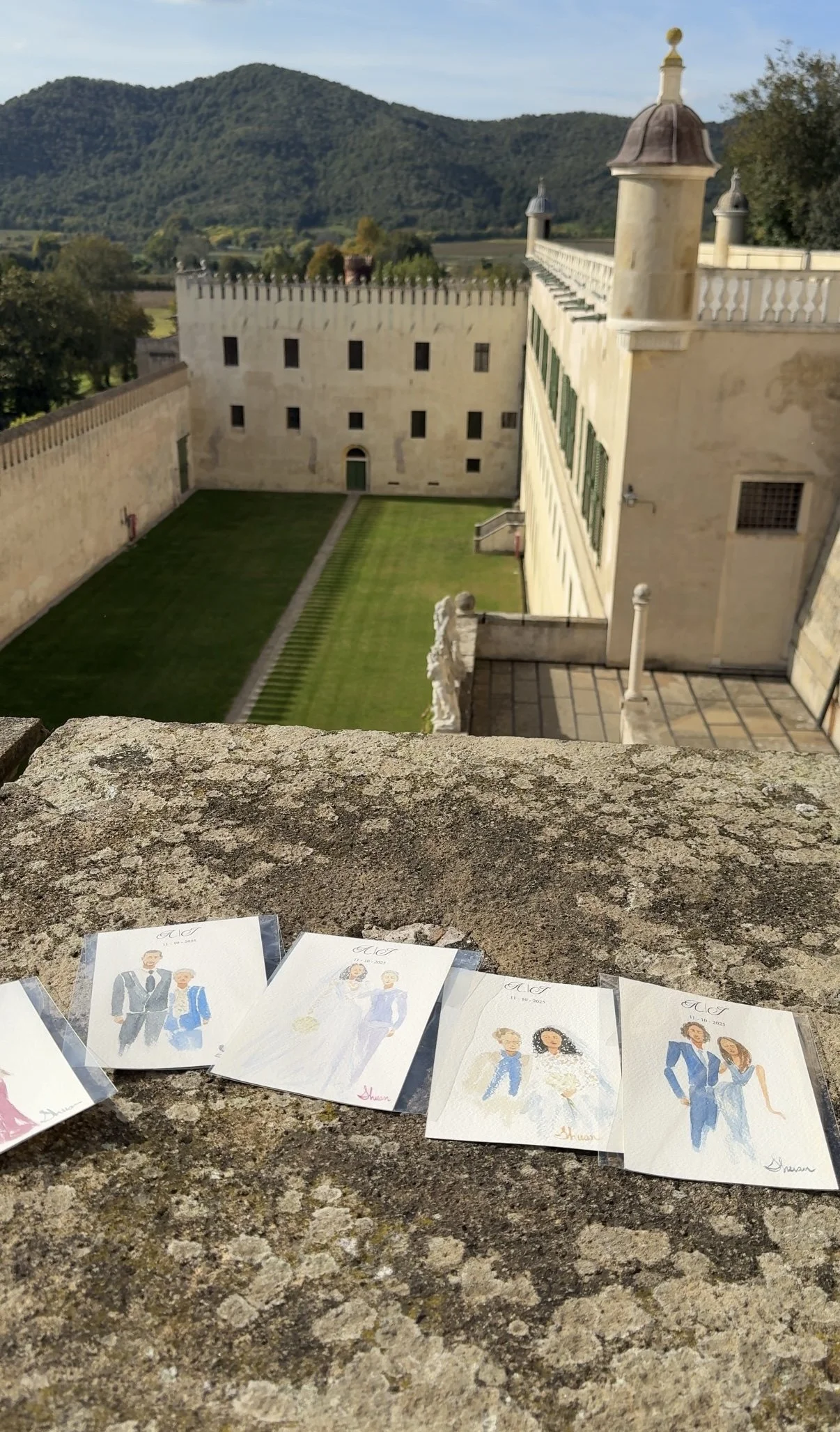A view from a stone balcony showing a castle courtyard with green grass, stone walls, and a building with green shutters against a backdrop of mountains and trees. There are Italy live wedding paintings of guests laid out on the stone surface in the 