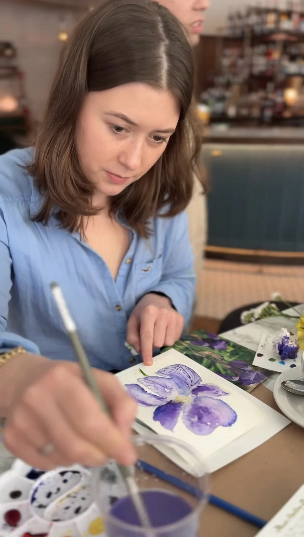 Woman sitting at a table in Italy painting a purple flower at a private watercolor workshop in Florence, with art supplies and photographs on the table.