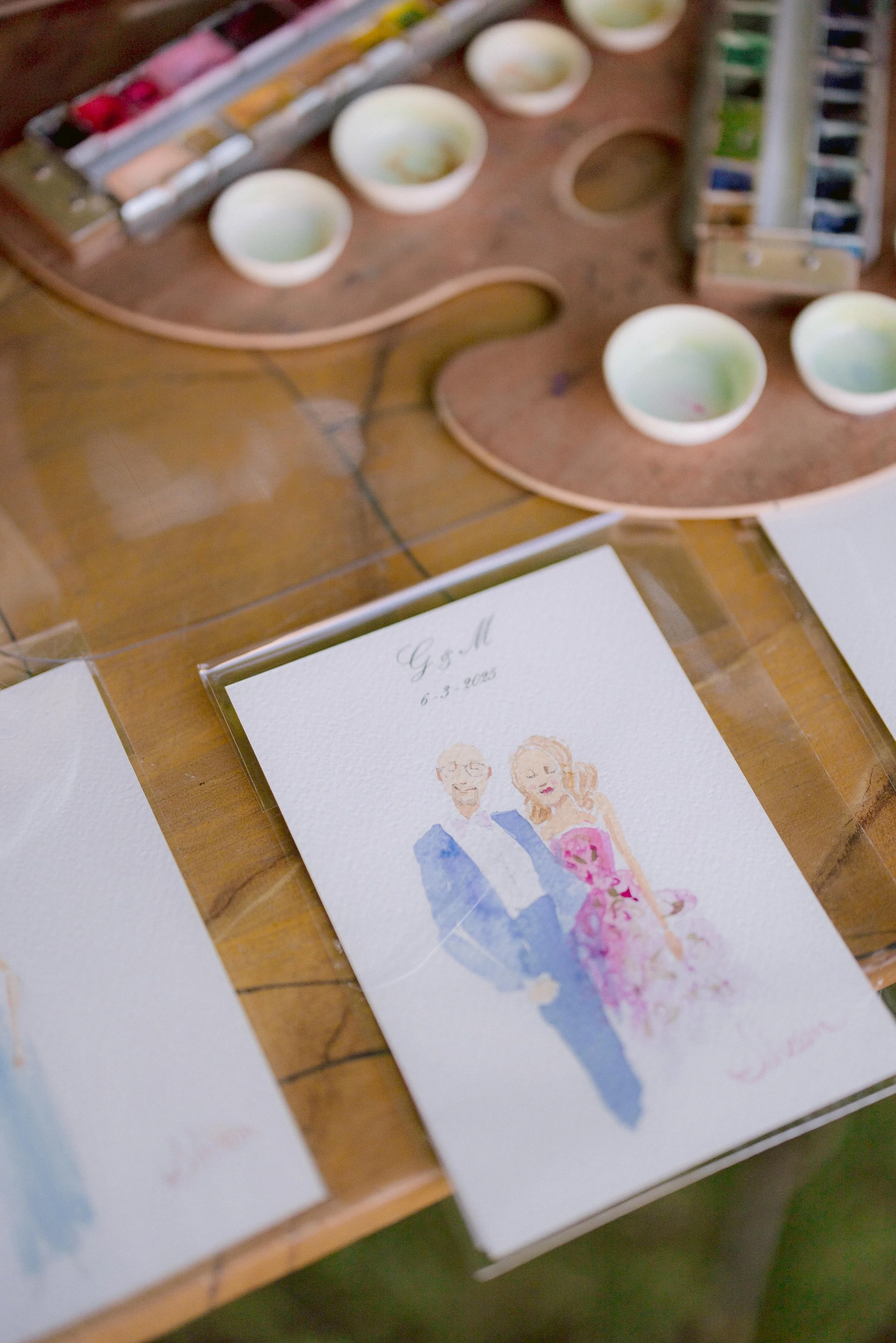An italy live wedding portrait featuring an illustrated couple, with the date 6.3.2025, placed on a wooden table. Surrounding the portrait are small bowls of watercolor paint and a palette, possibly for creating or decorating the painting.