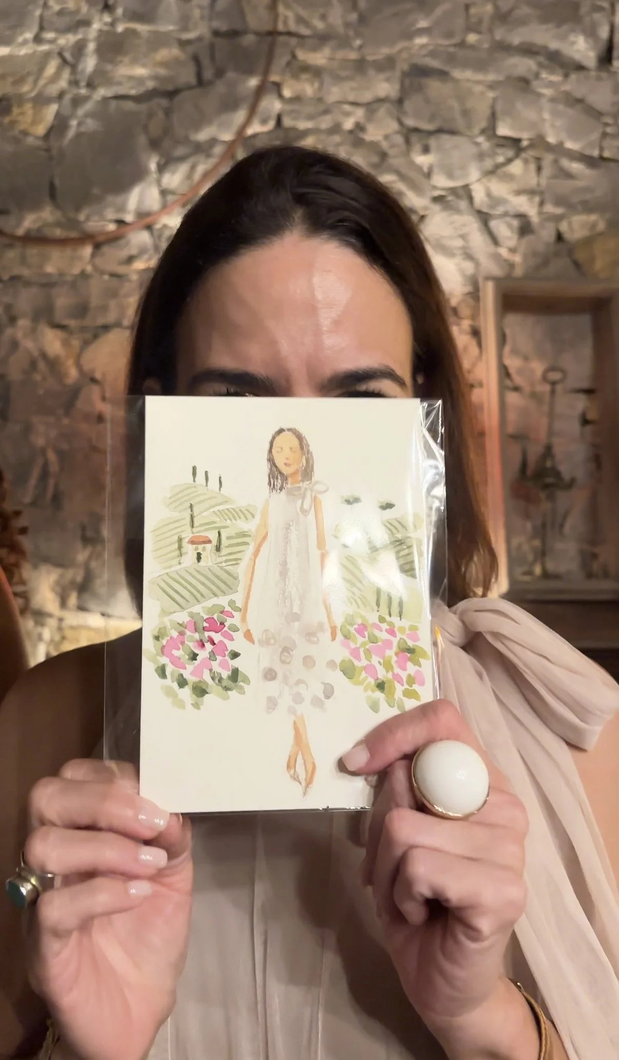 Woman holding an Italy watercolor portrait illustration of a woman standing in a field of flowers, partially covering her face, in front of a stone wall.
