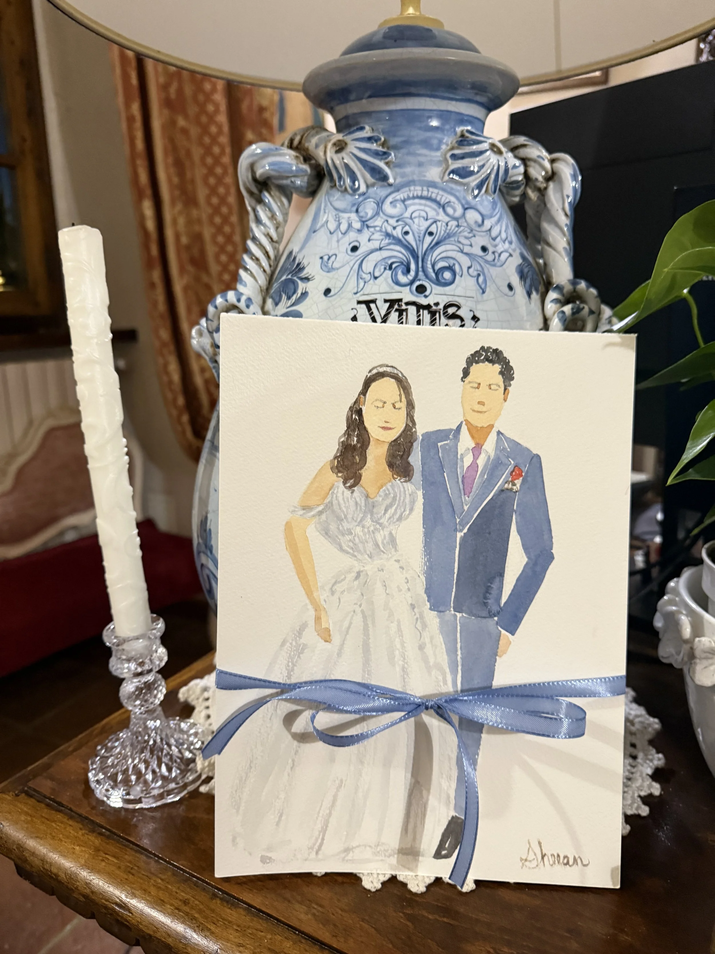  Italy live wedding watercolor illustration of a bride in a white dress and a groom in a dark blue suit. The card is tied with a blue ribbon and has a signature at the bottom right. It is placed on a wooden table with a white lace doily, next to a ta