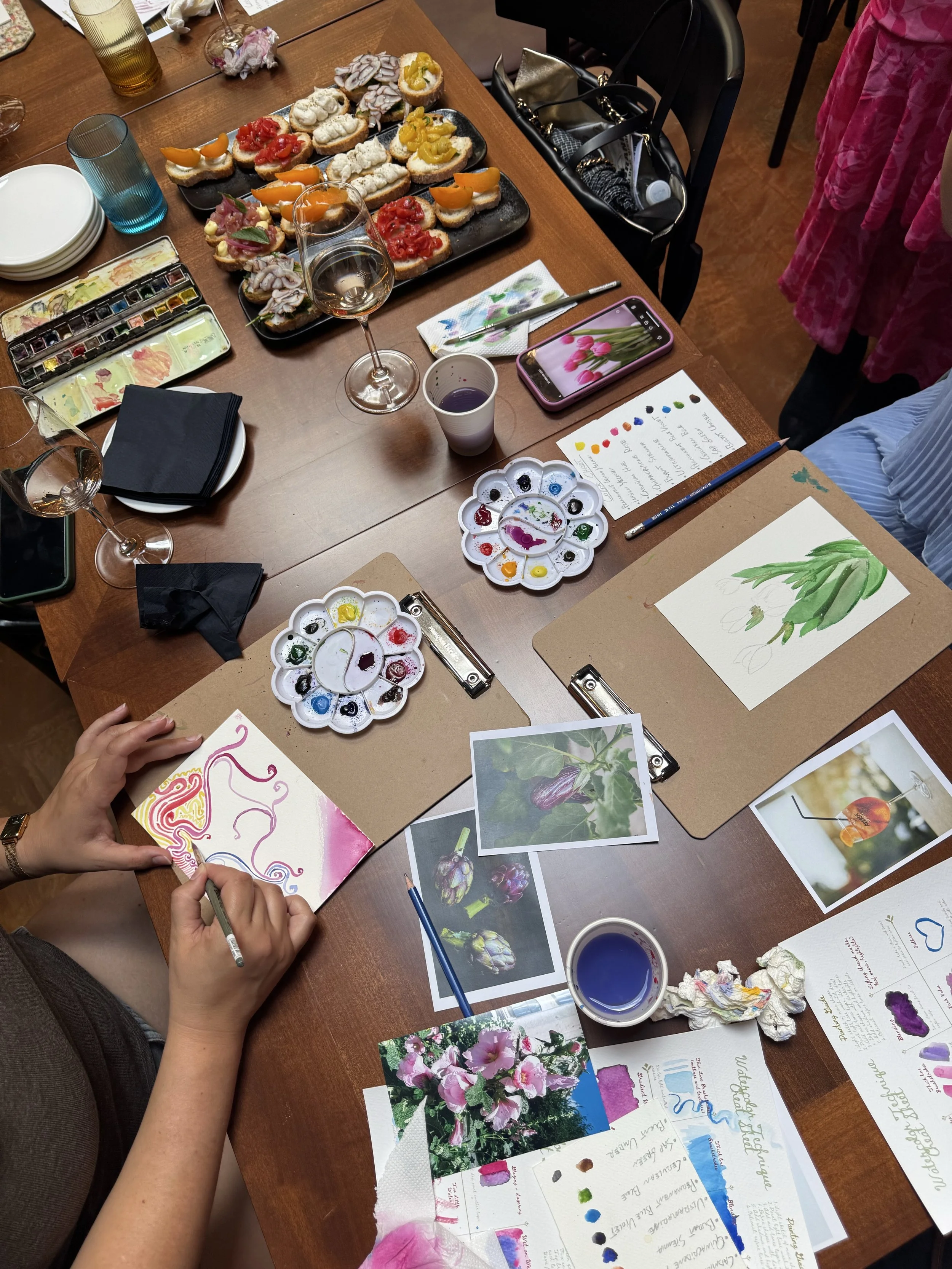 People participating in a flower painting or botanical art class in Florence Italy with watercolor paints, photographs of plants, and a table filled with snacks, drinks, and art supplies.