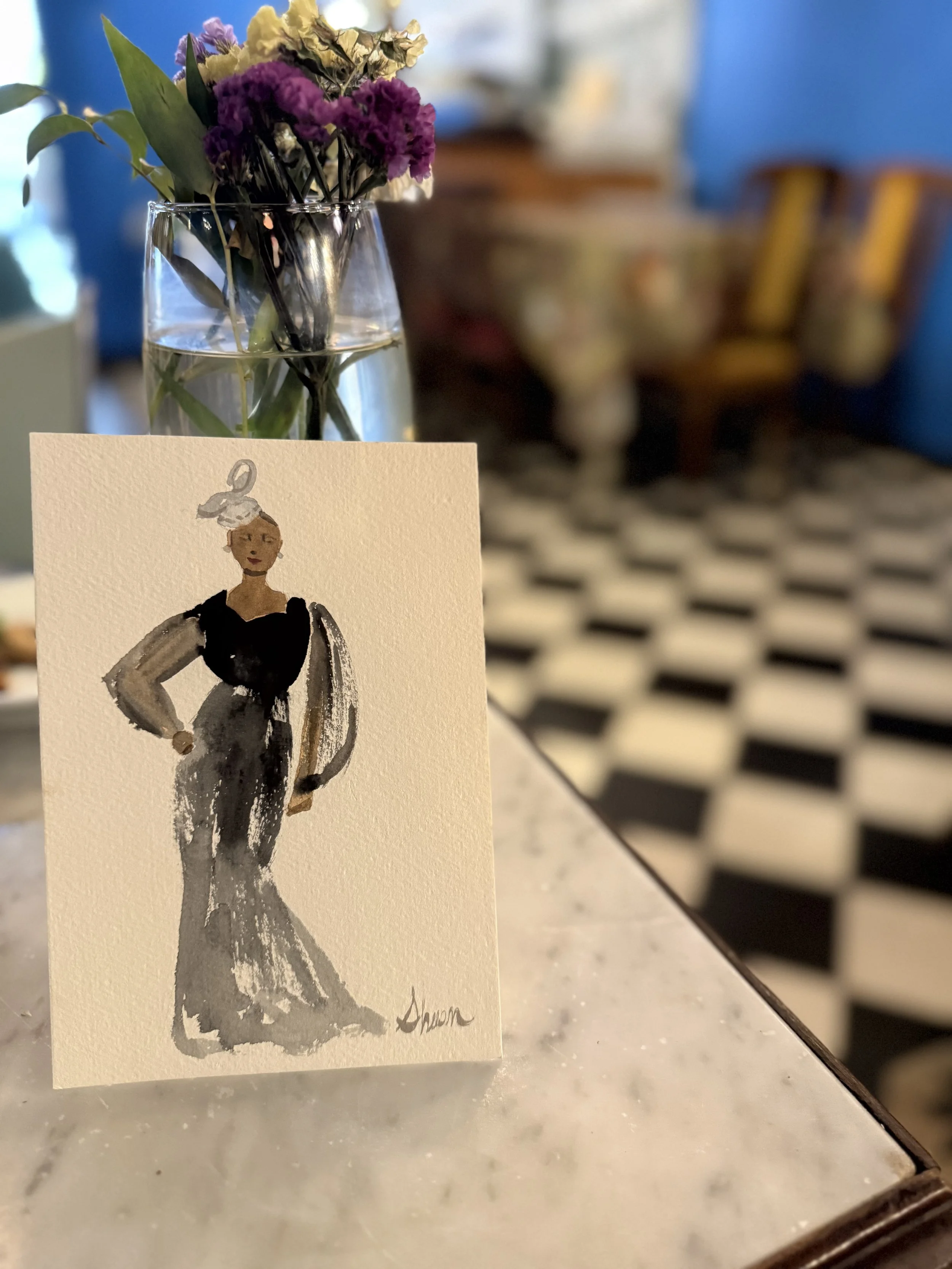 Italy live wedding guest portrait of a woman wearing a long, black and gray dress with a jacket, standing confidently with one hand on her hip. Behind her are some drawn white angel wings. The illustration appears on a small greeting card placed on a
