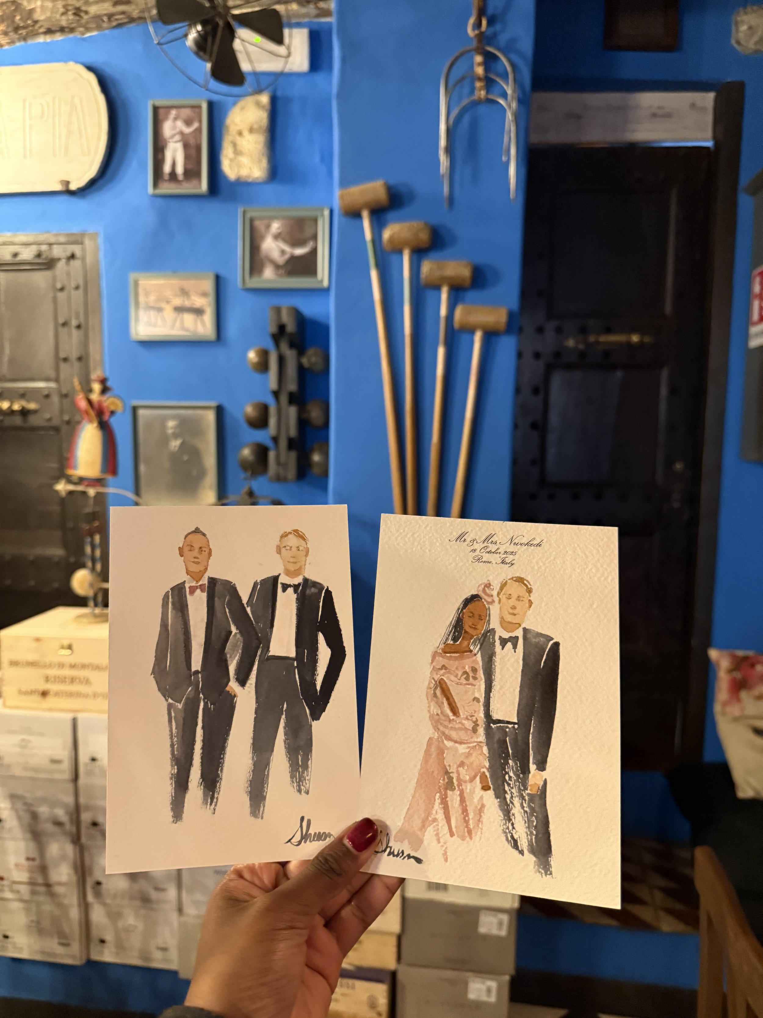 A person holding two live wedding guest portraits in Italy in front of a blue wall decorated with pictures, tools, and vintage items in a room with wooden furniture.