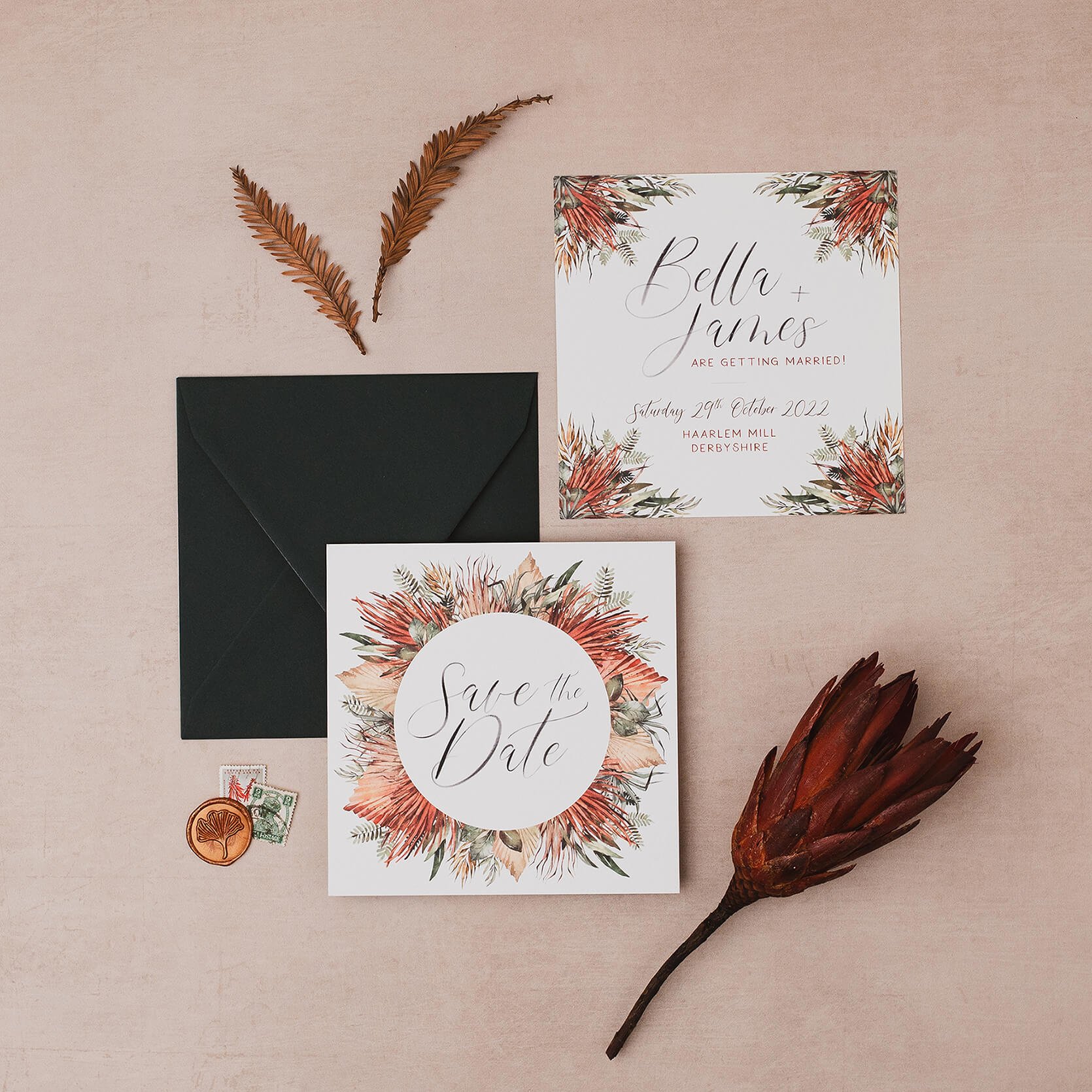 Wedding invitation and save the date cards with floral designs, an black envelope, dried leaves, and a large dried flower on a light pink background.