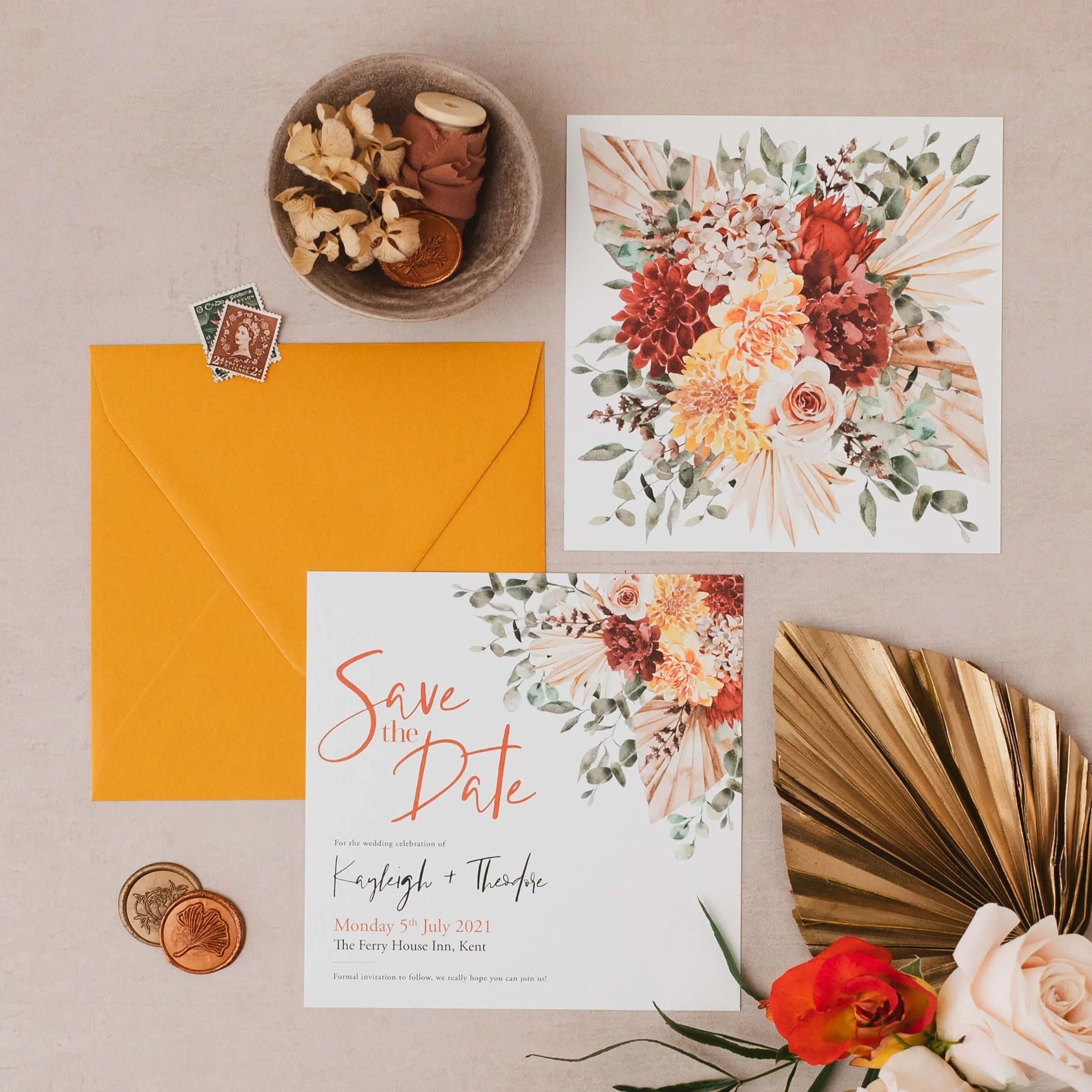 Wedding invitation with floral design, yellow envelope, vintage stamps, gold decorative fan, and a bowl with dried flowers, candles, and a copper coin.