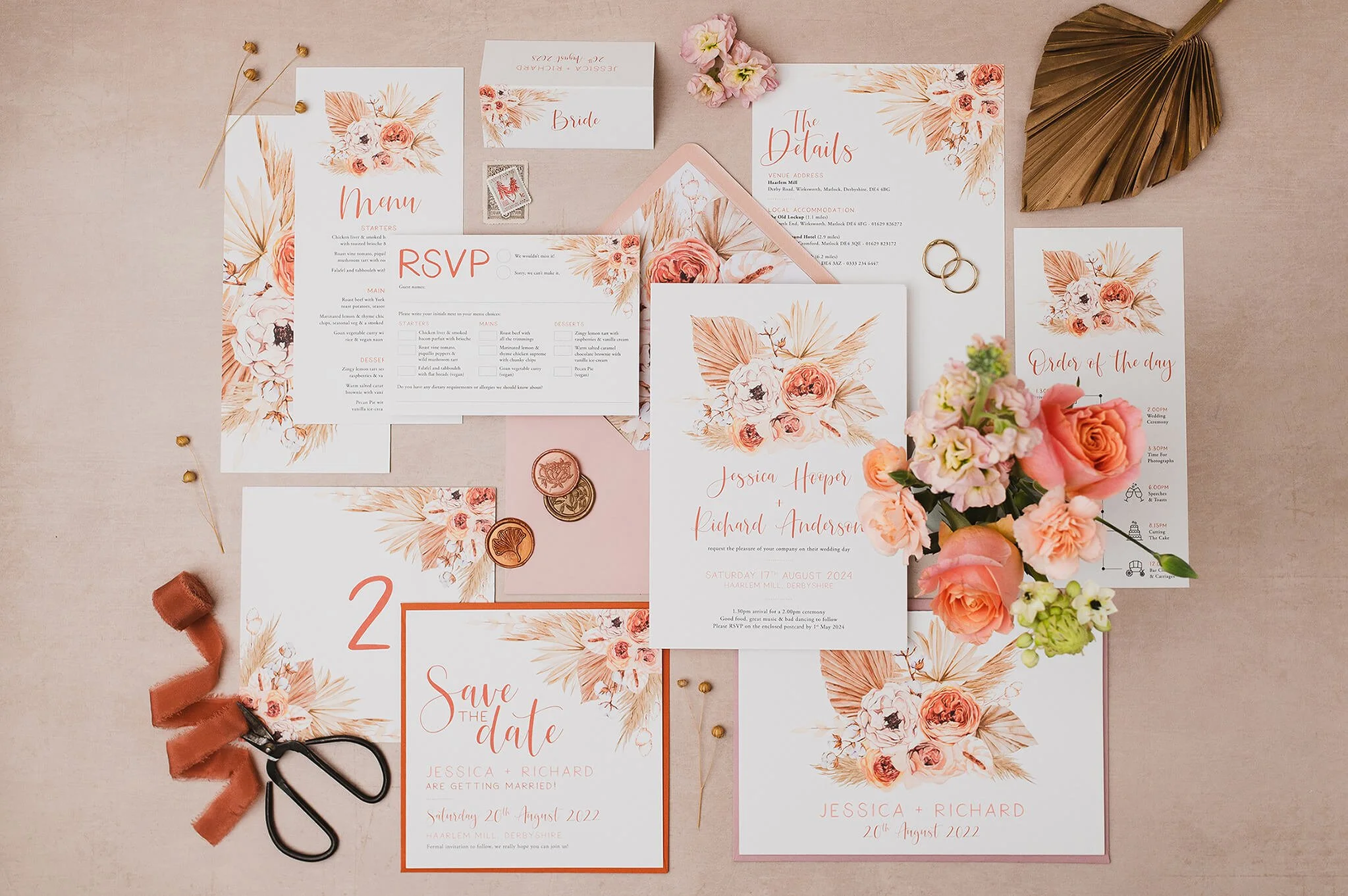 Wedding stationery set including save-the-date and pampas wedding invitations with a floral design of pink and cream roses, dried leaves, and beige palm leaves, laid out on a beige surface.