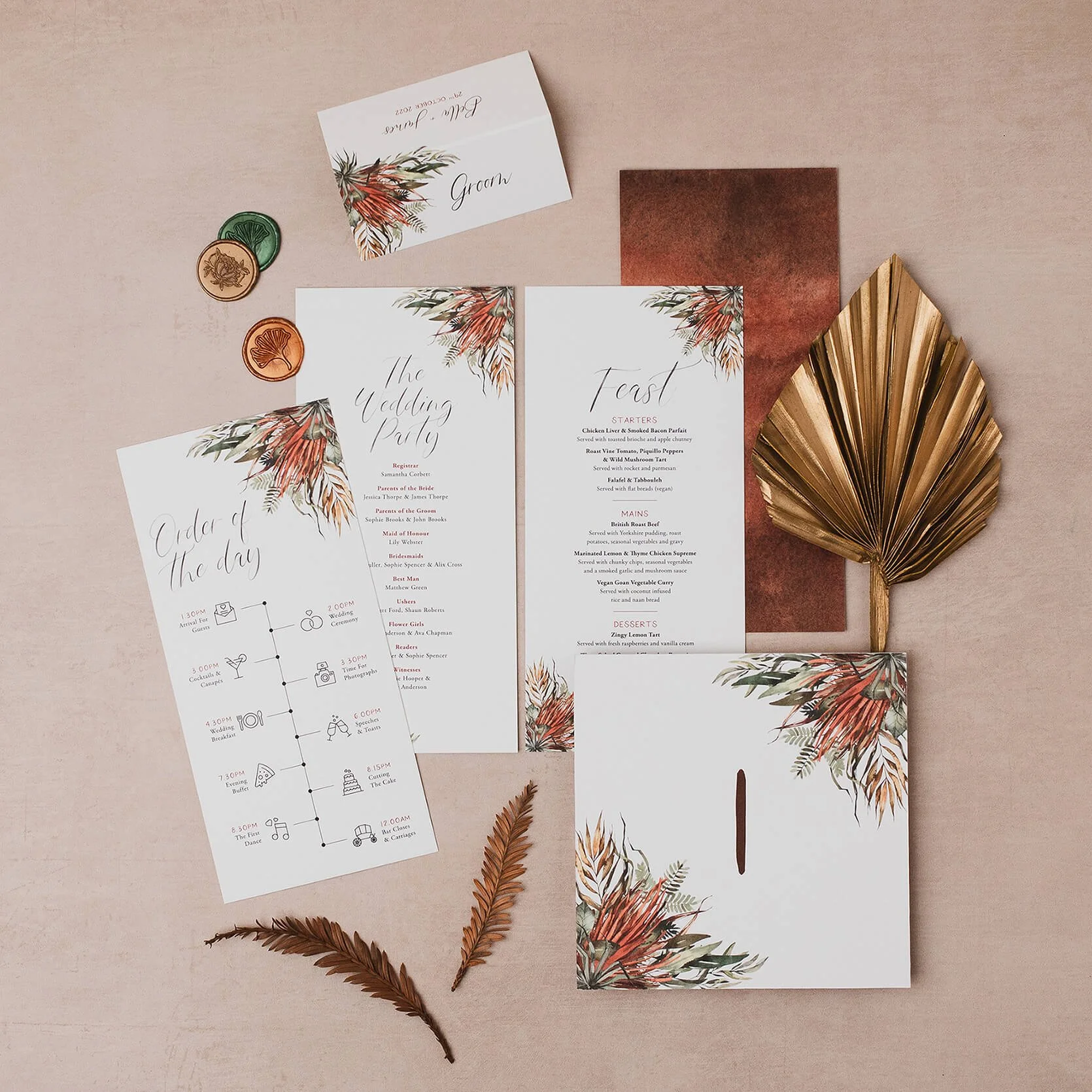 A wedding décor setup featuring wedding invitations with floral designs, a place card labeled 'Groom', a menu, an order of the day schedule, and envelopes, all arranged on a pink background with decorative dried leaves and a golden palm leaf.