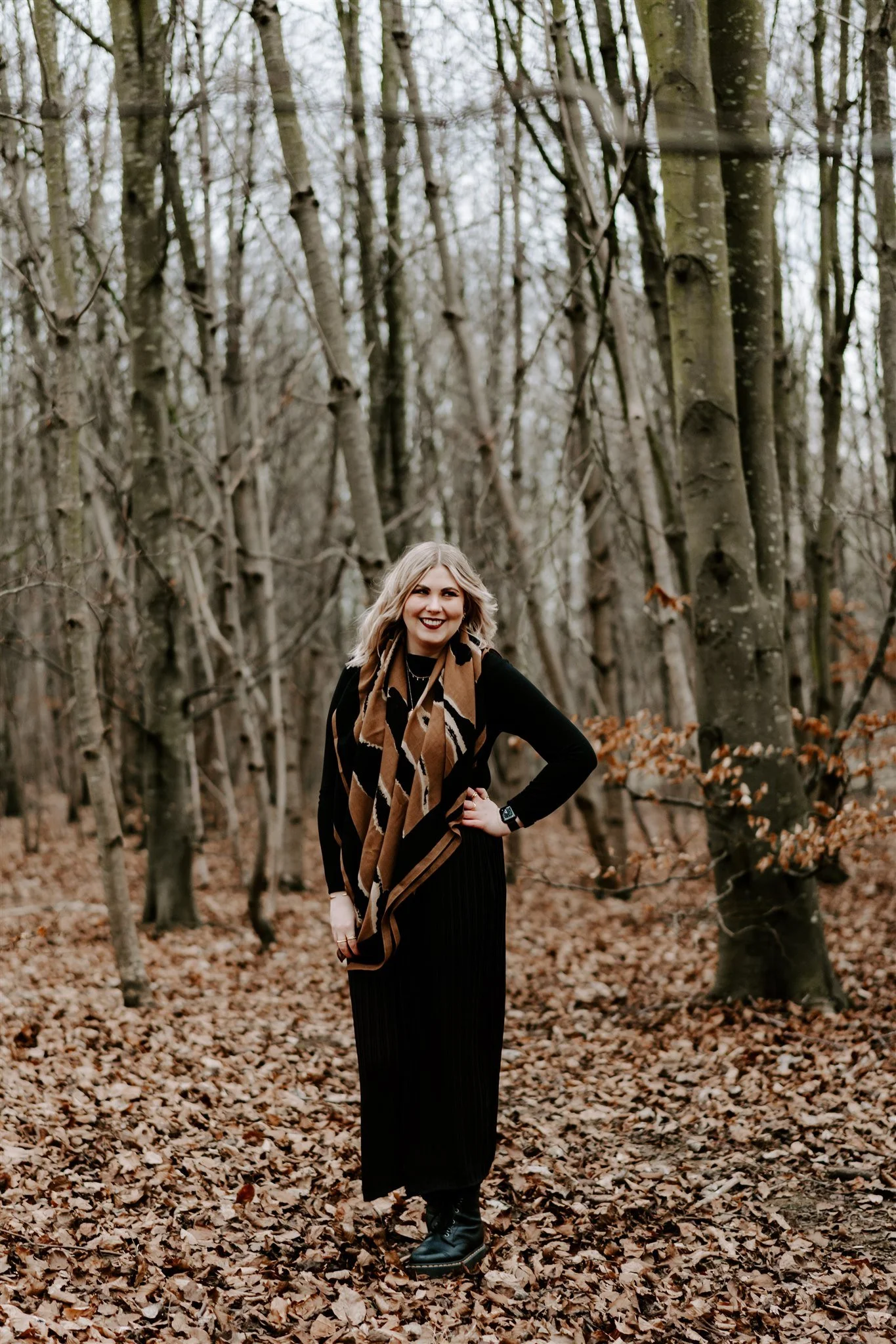 A woman with blonde hair wearing a black long-sleeve shirt, black pants, and a brown patterned scarf standing on a leaf-covered forest path, smiling, with trees in the background during autumn.
