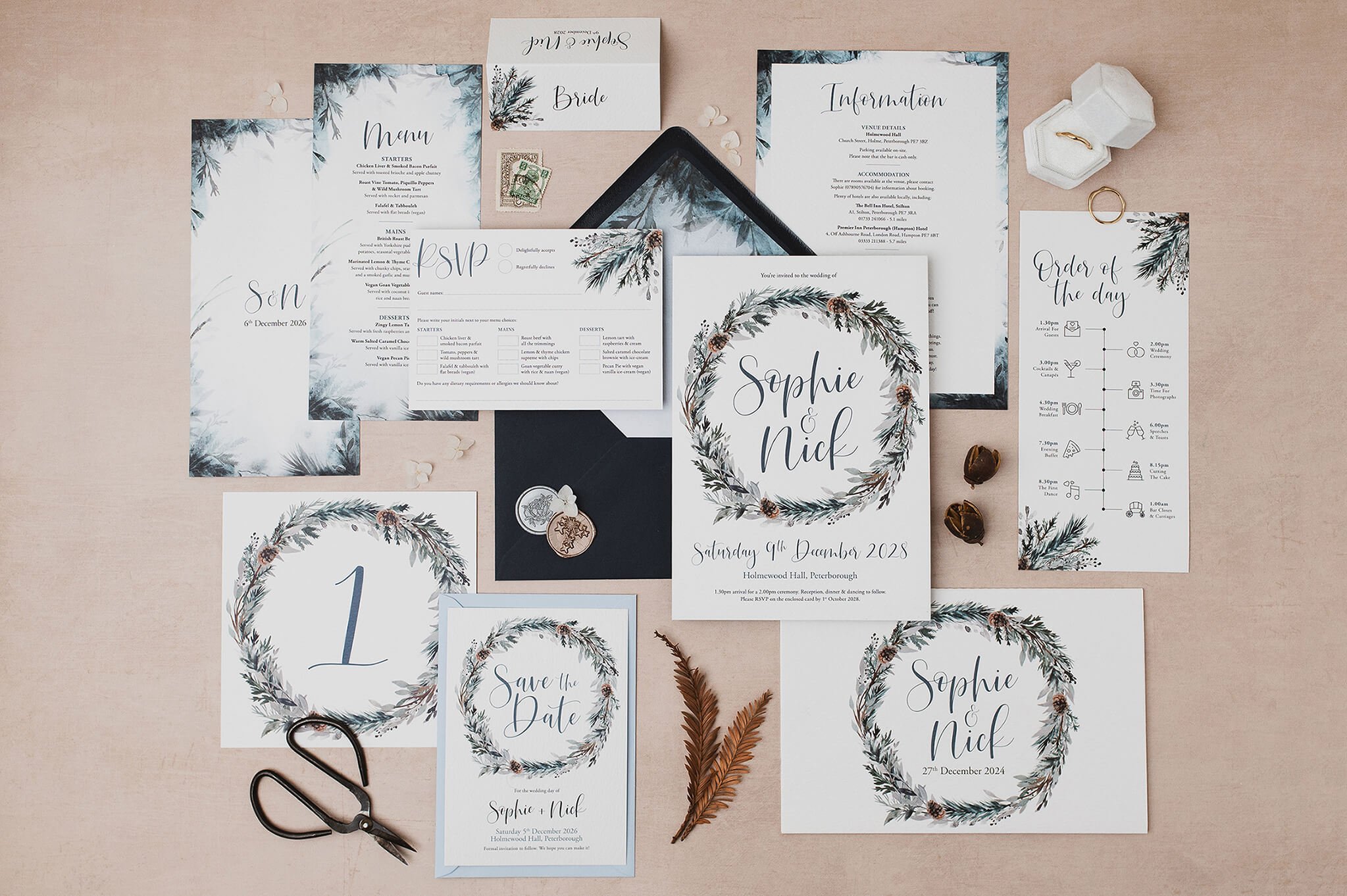 Set of navy blue toned wedding stationery including a save the date card, winter wedding invitation, menu, RSVP card, and order of service, all featuring a winter foliage wreath design with pine cones and muted green and gray leaves.