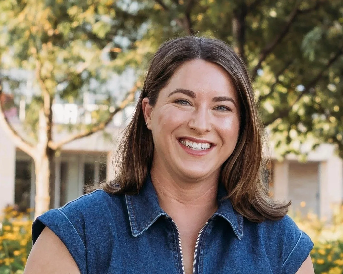 A woman with shoulder-length brown hair smiling outdoors, wearing a sleeveless blue denim shirt, with trees and buildings in the background.