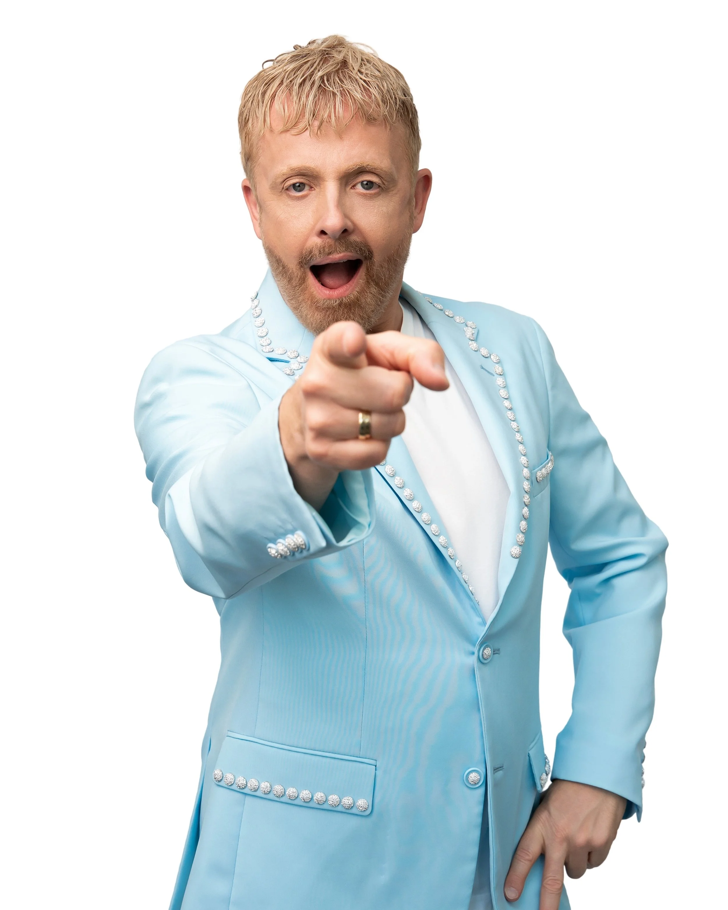 Ein lächelnder Mann mit Bart trägt einen pinken Anzug mit Perlen, eine hellblaue Hemd und eine grüne Schleife, steht vor einem weißen Hintergrund.